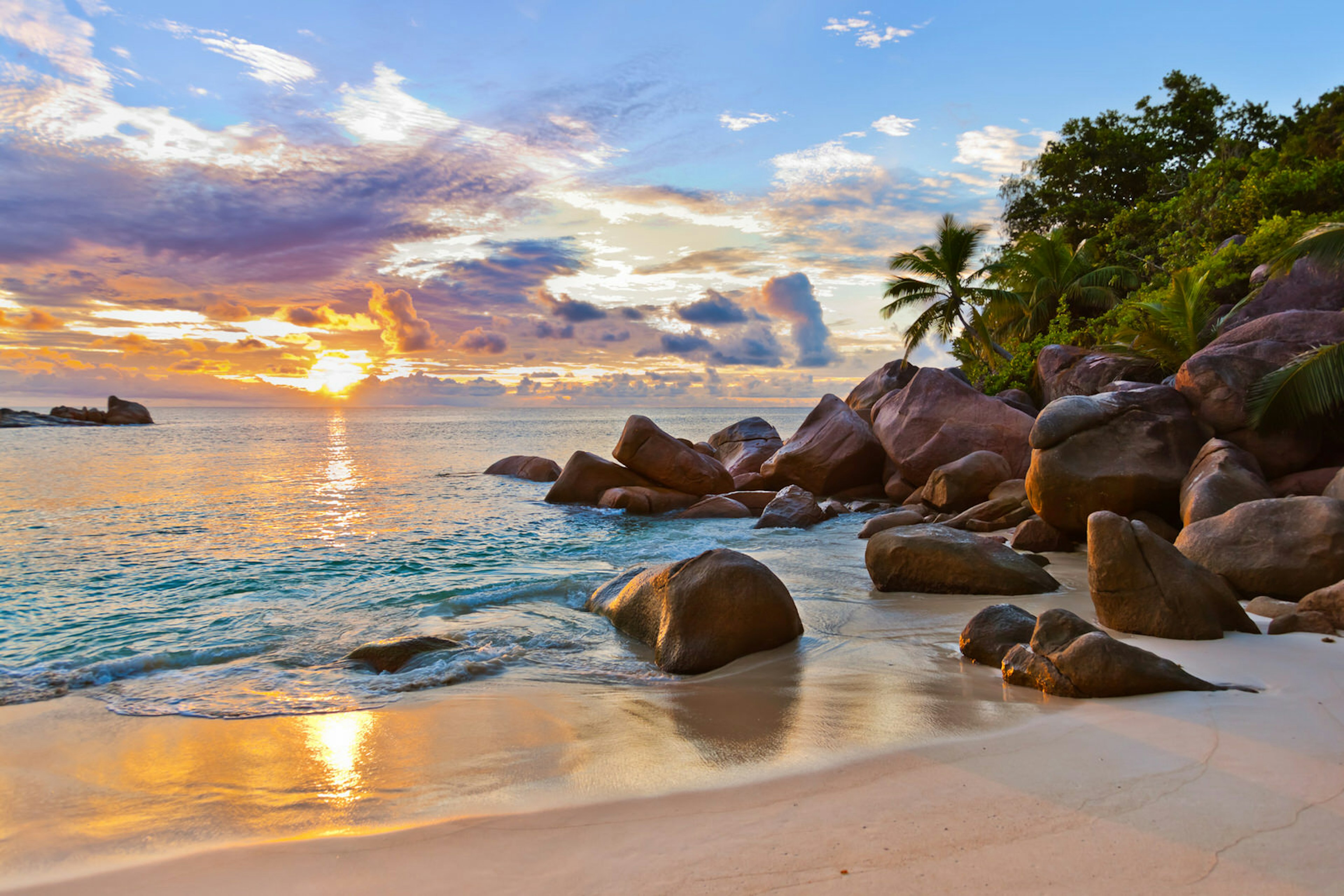 A beautiful cove in the Seychelles; the sky is a dramatic canvas of clouds and the setting sun, while the sea is greeny-blue and surf laps at a golden shore; at the end of the beach, large grey rocks rise to meet palm trees and other greenery.