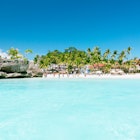 Boracay's White Beach viewed from a boat is wedged between an azure sky and clear, turquoise sea. Behind the sand there is an abundance of green palm trees, a handful of beach bars and some people enjoying the beach