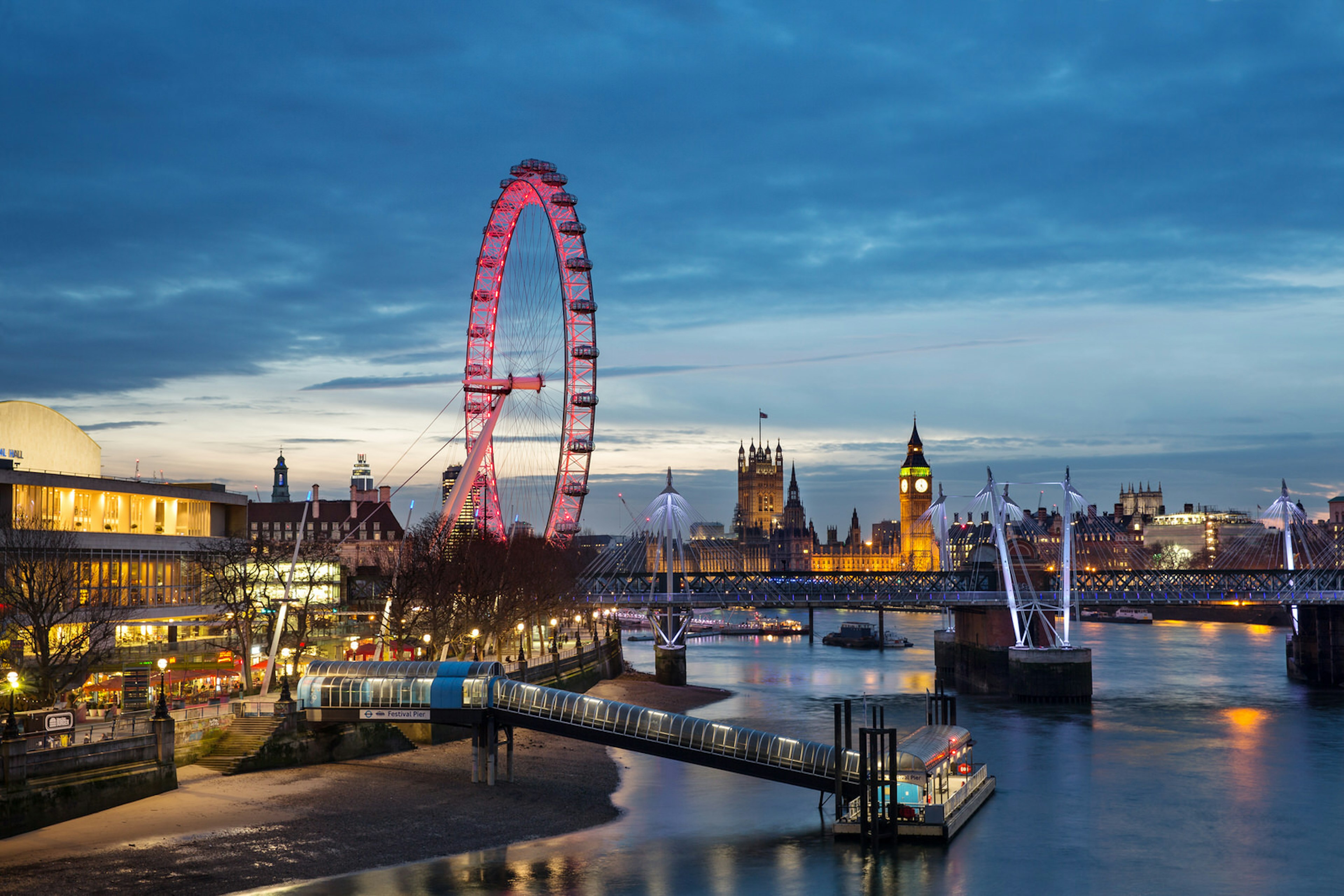 The London Eye is lit up red, the Thames is at low tide and in the background, the Palace of Westminster is lit up against a moody sky