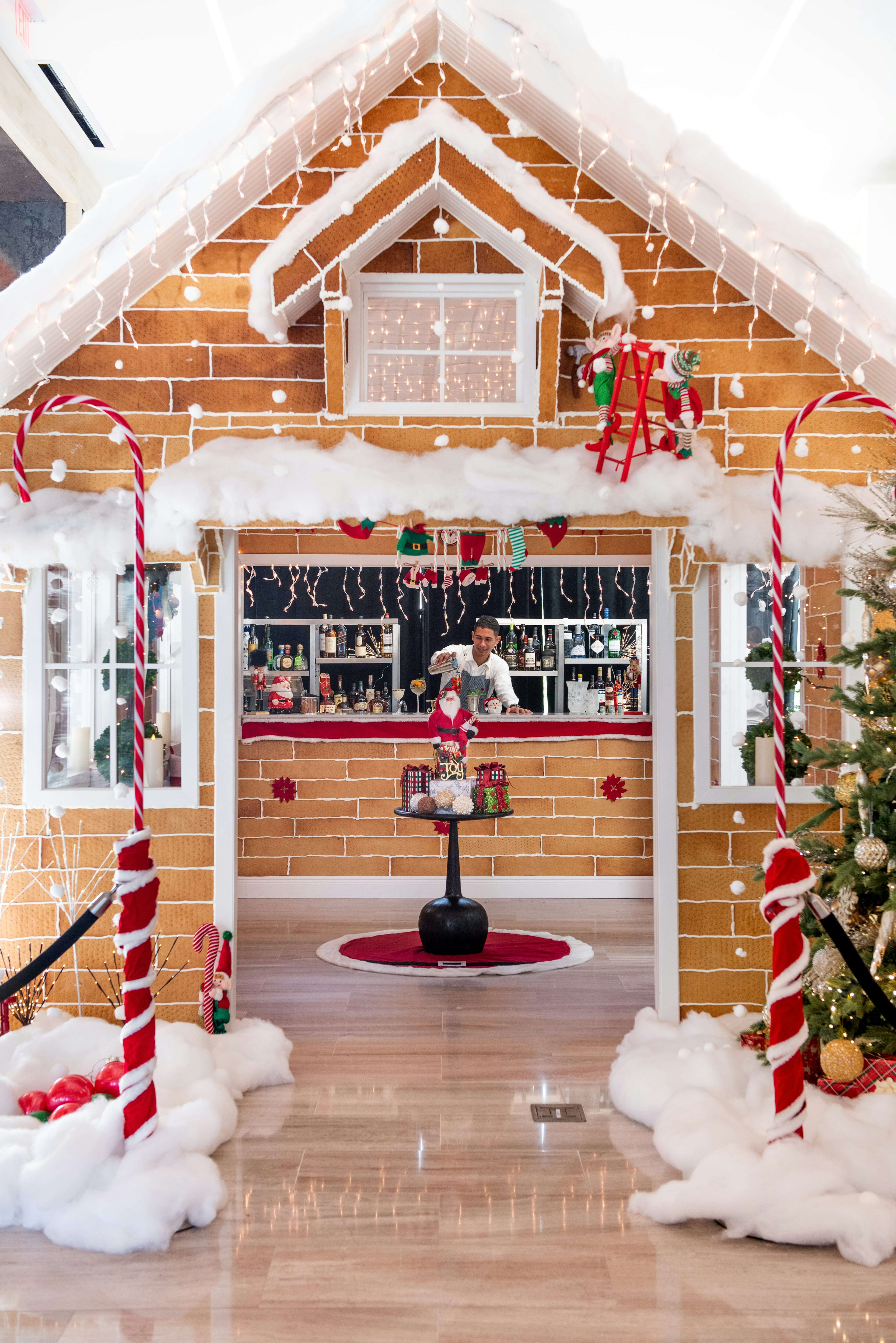 A life-size gingerbread house at Kimpton Seafire Resort + Spa; the house is a bar, and has been decorated with candy canes and fake snow. Through the door the bartender can be seen making cocktails.