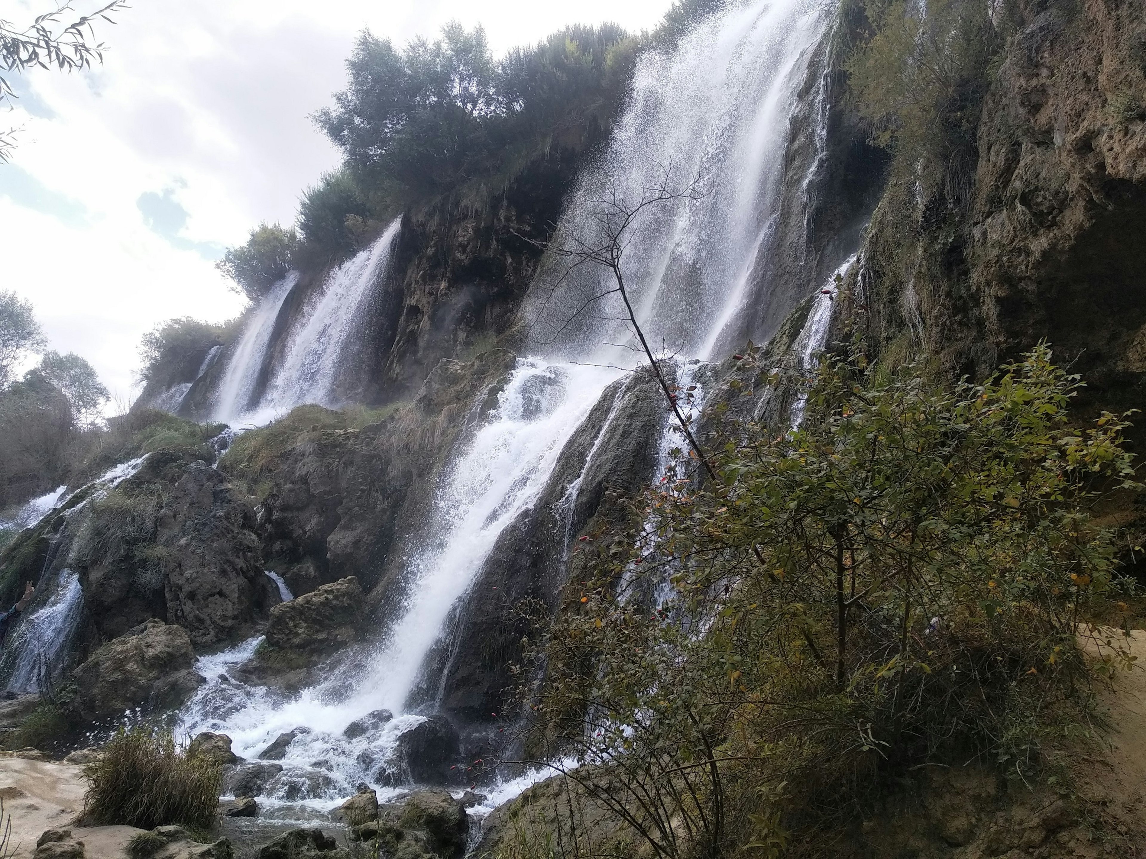 Waterfalls cascade down a rock face, with several bushes and trees either side