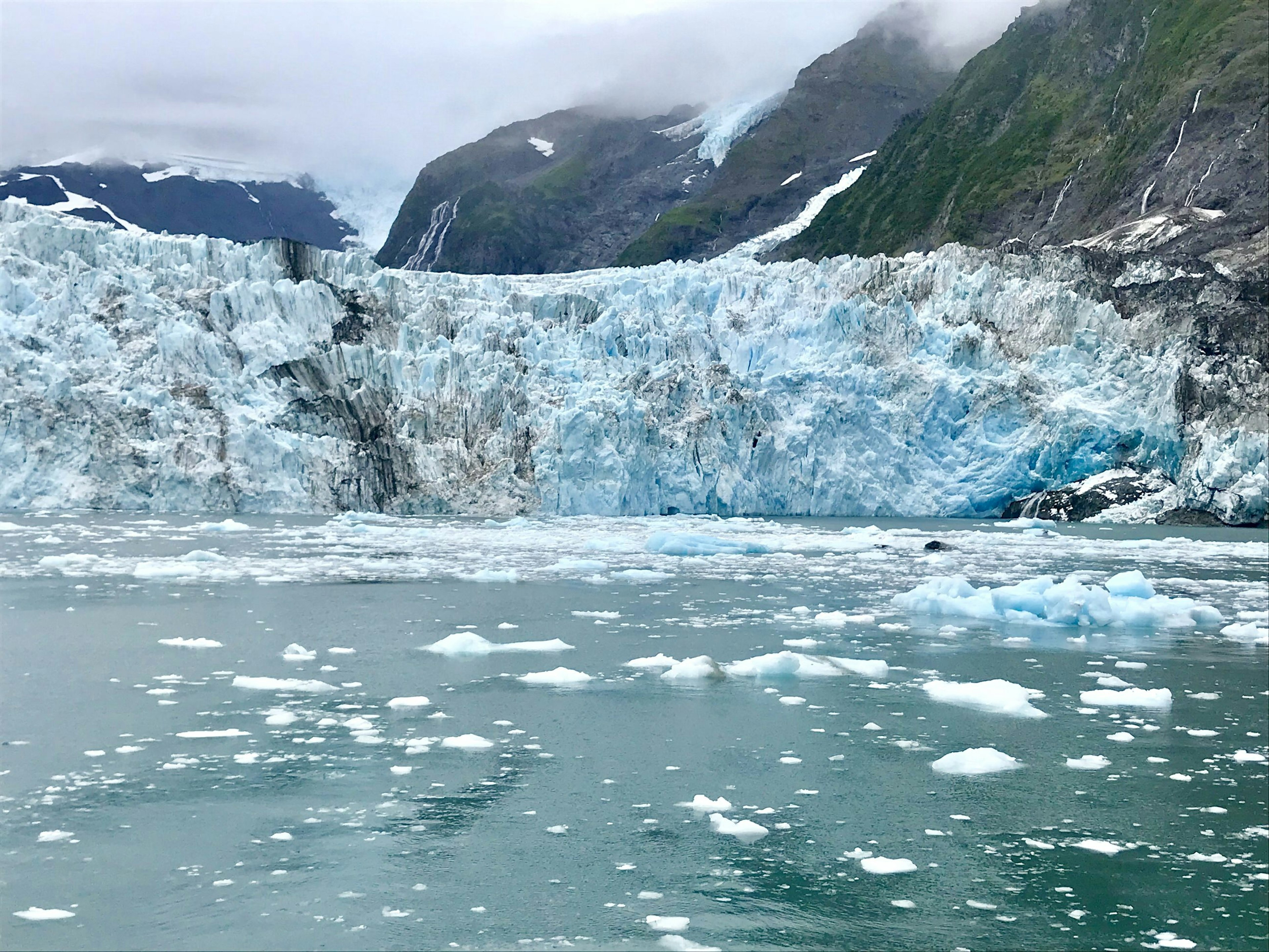 Chunks of ice floating in the ice-blue water beneath a calving glacier. Mist lingers  around the mountains above.