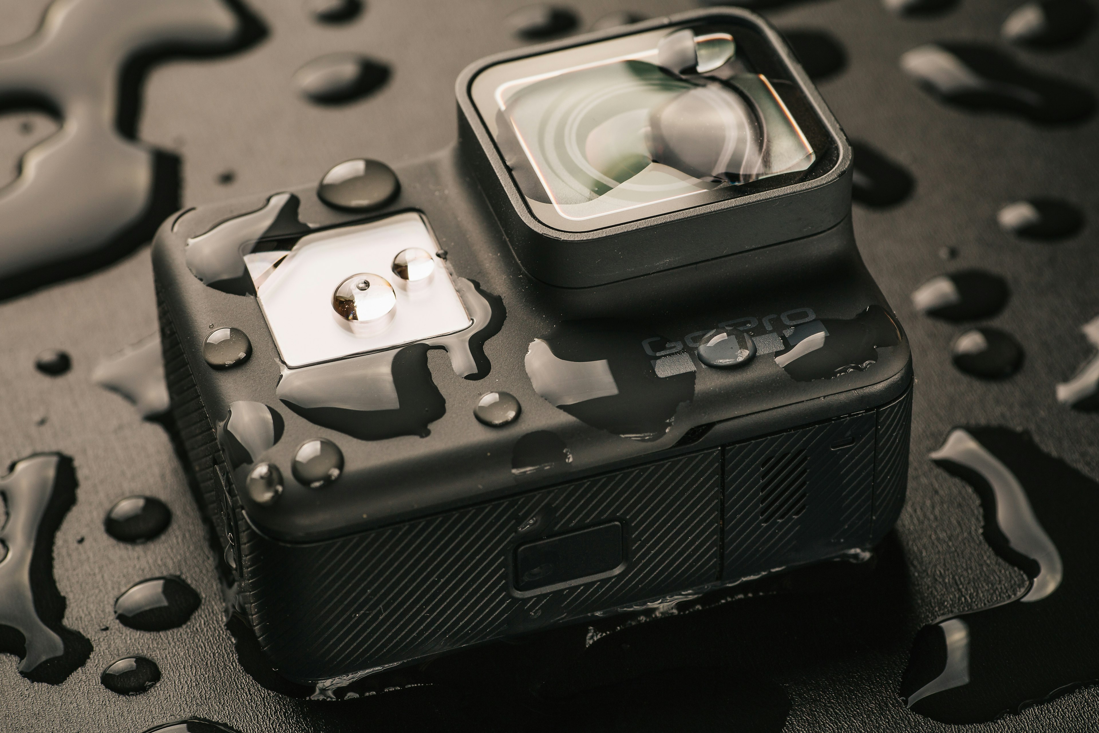 A GoPro Camera is on a black surface, covered in globules of water.