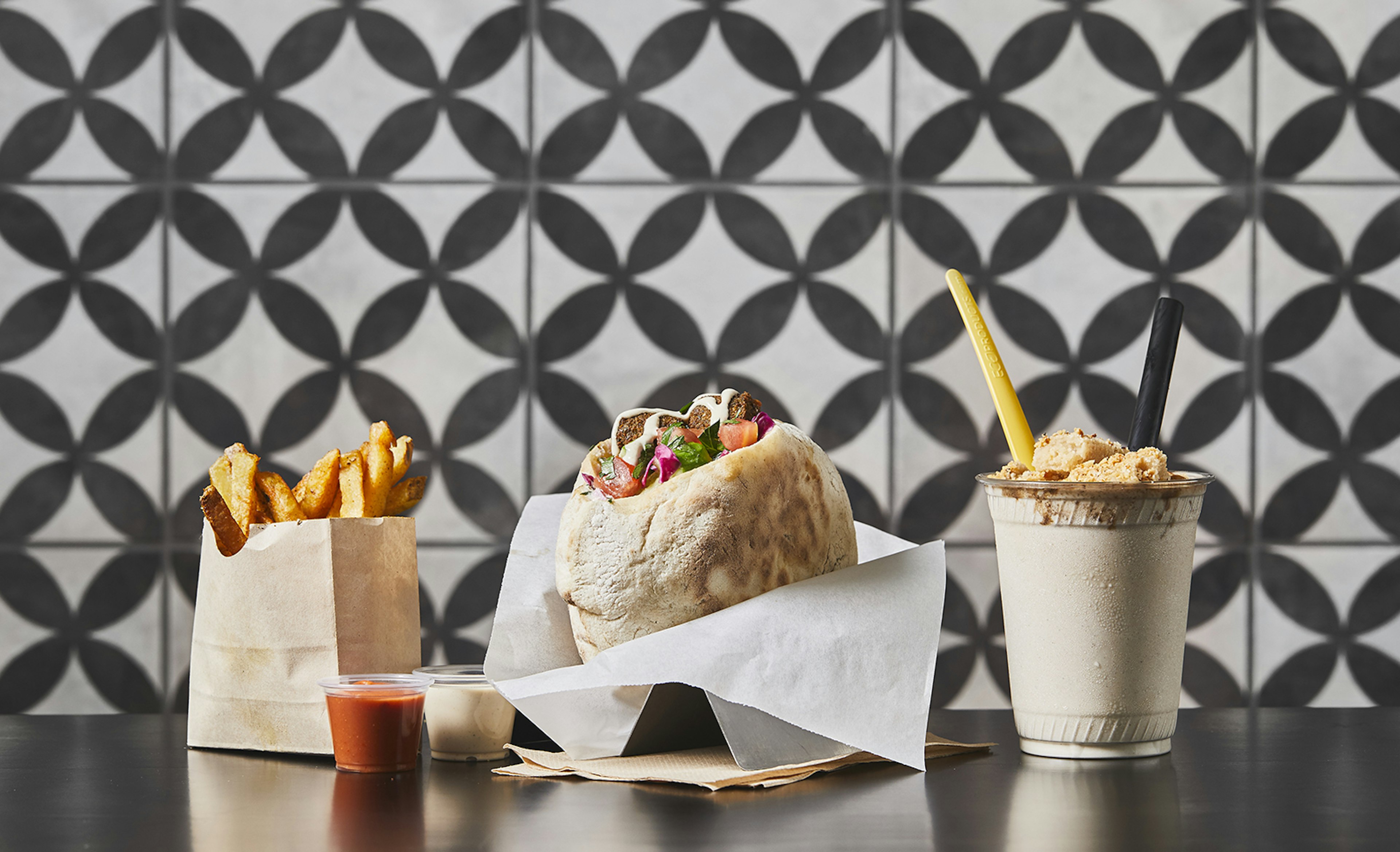 Golden fries in a paper bag, a falafel in a wax-paper wrapper and a frosty milkshake with a yellow straw sit on a counter in front of a black-and-white tile background at Goldie's in Philadelphia