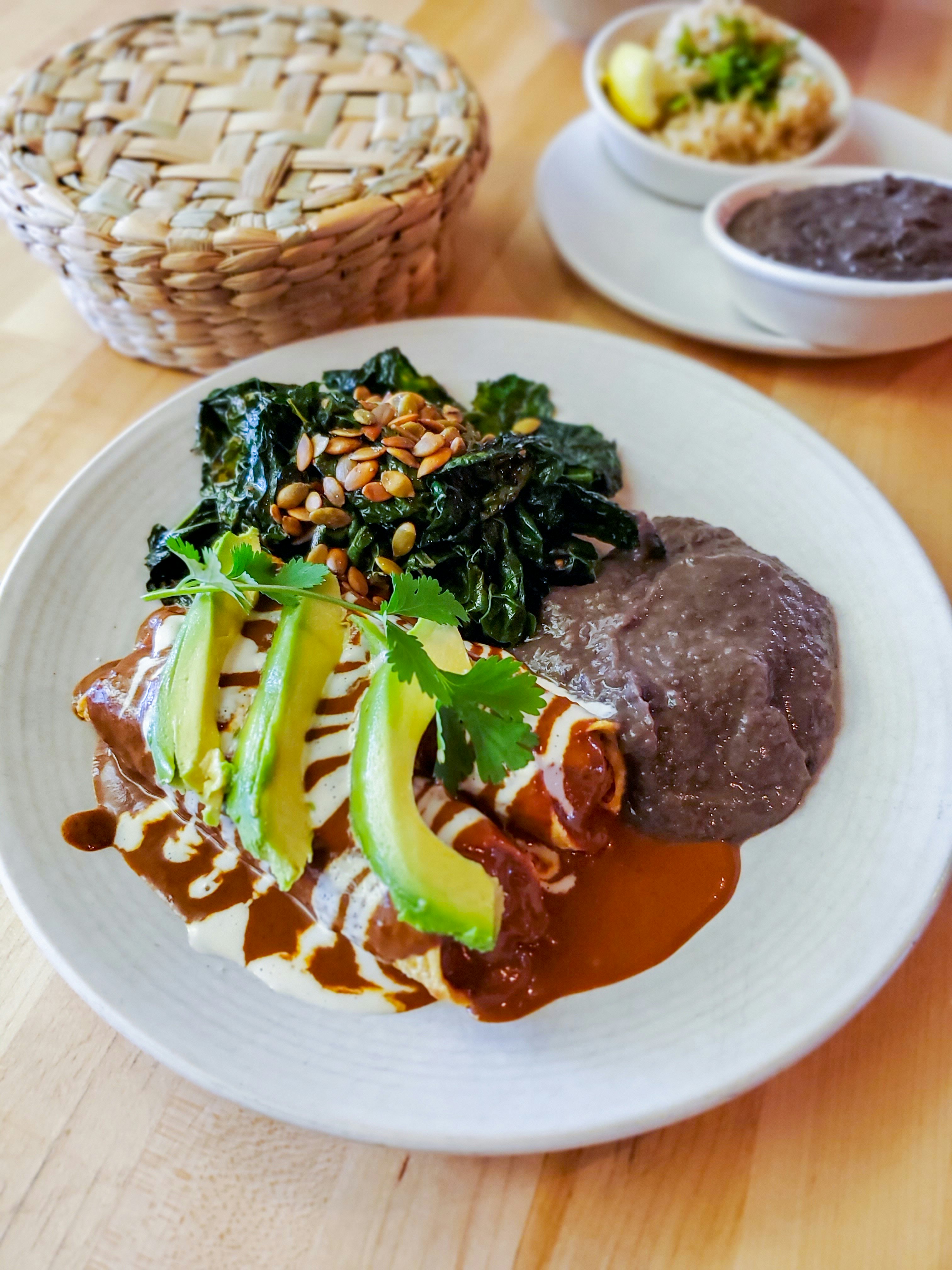 A plate filled with vegan enchiladas, sliced avocado, refried beans and greens topped with pine nuts; Bay Area vegan restaurant