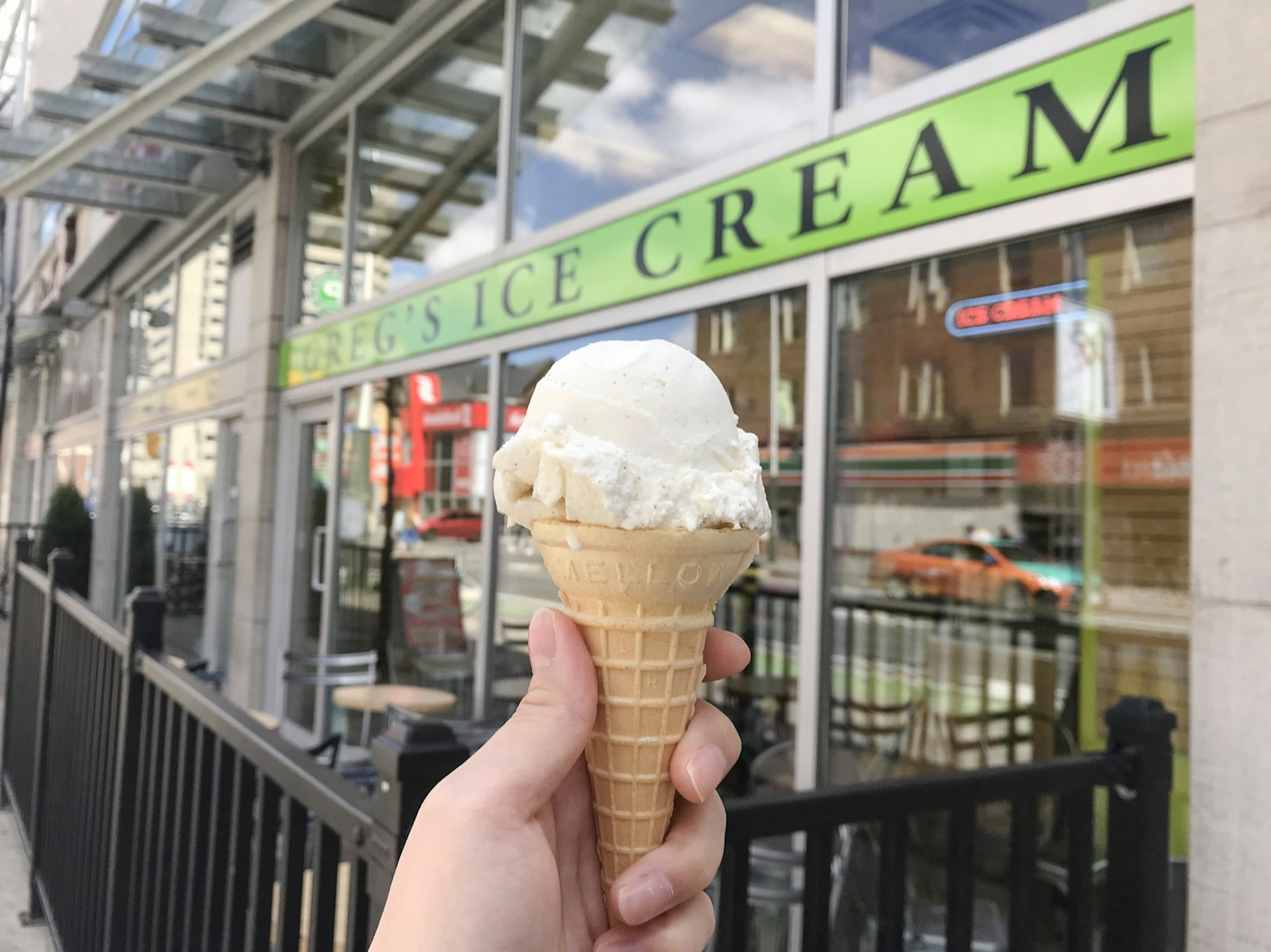 A hand holds a simple-looking ice cream cone in a plain sugar cone in front of a shop