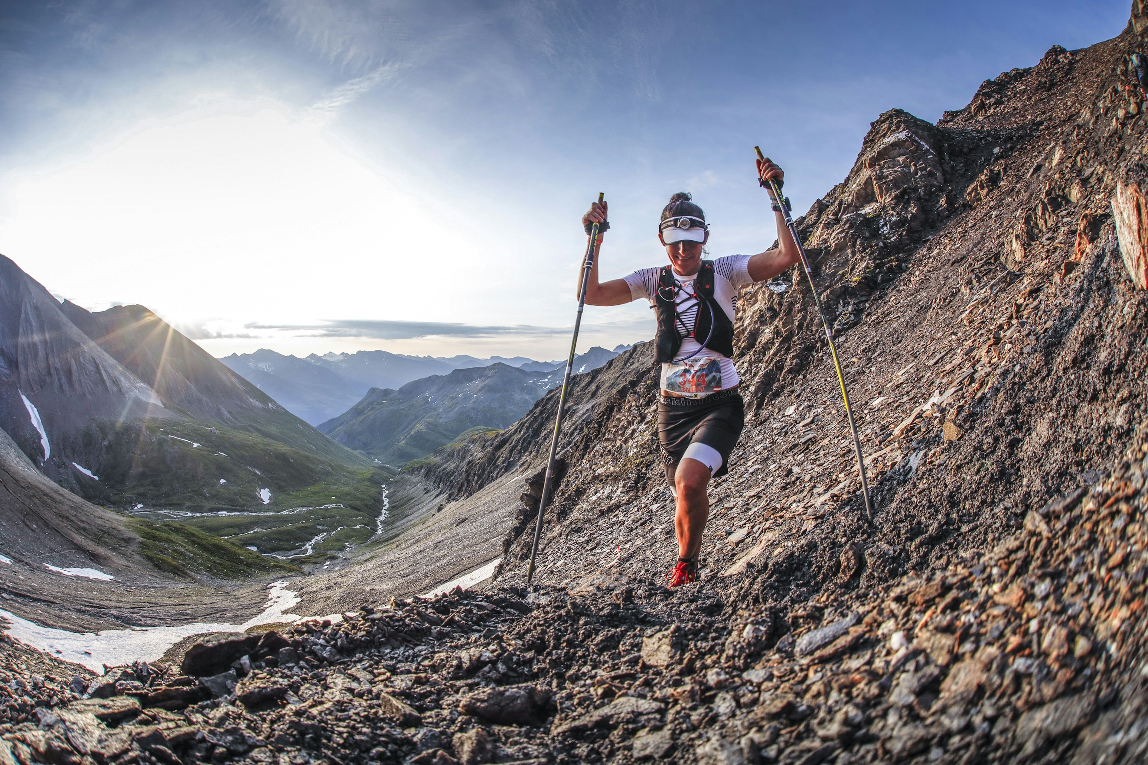 A female runner with two long poles ascends a scree slope towards the camera on the side of a dramatic valley; in the background the valley descends back towards the lowering sun.