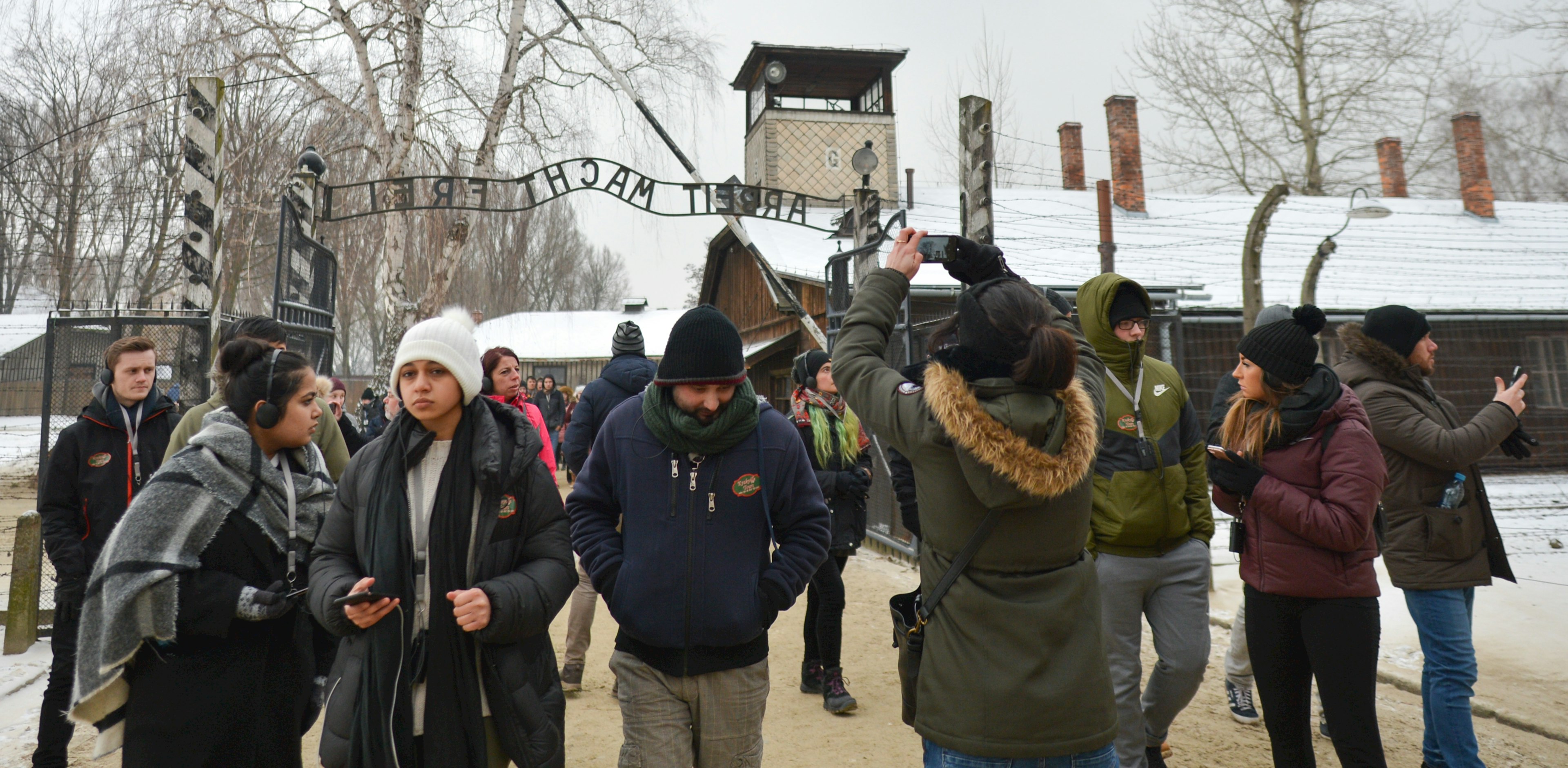 A group of schoolchildren stand inside the gates of Auschwitz. The famous Arbeit macht frei (work will set you free) sign is visible.