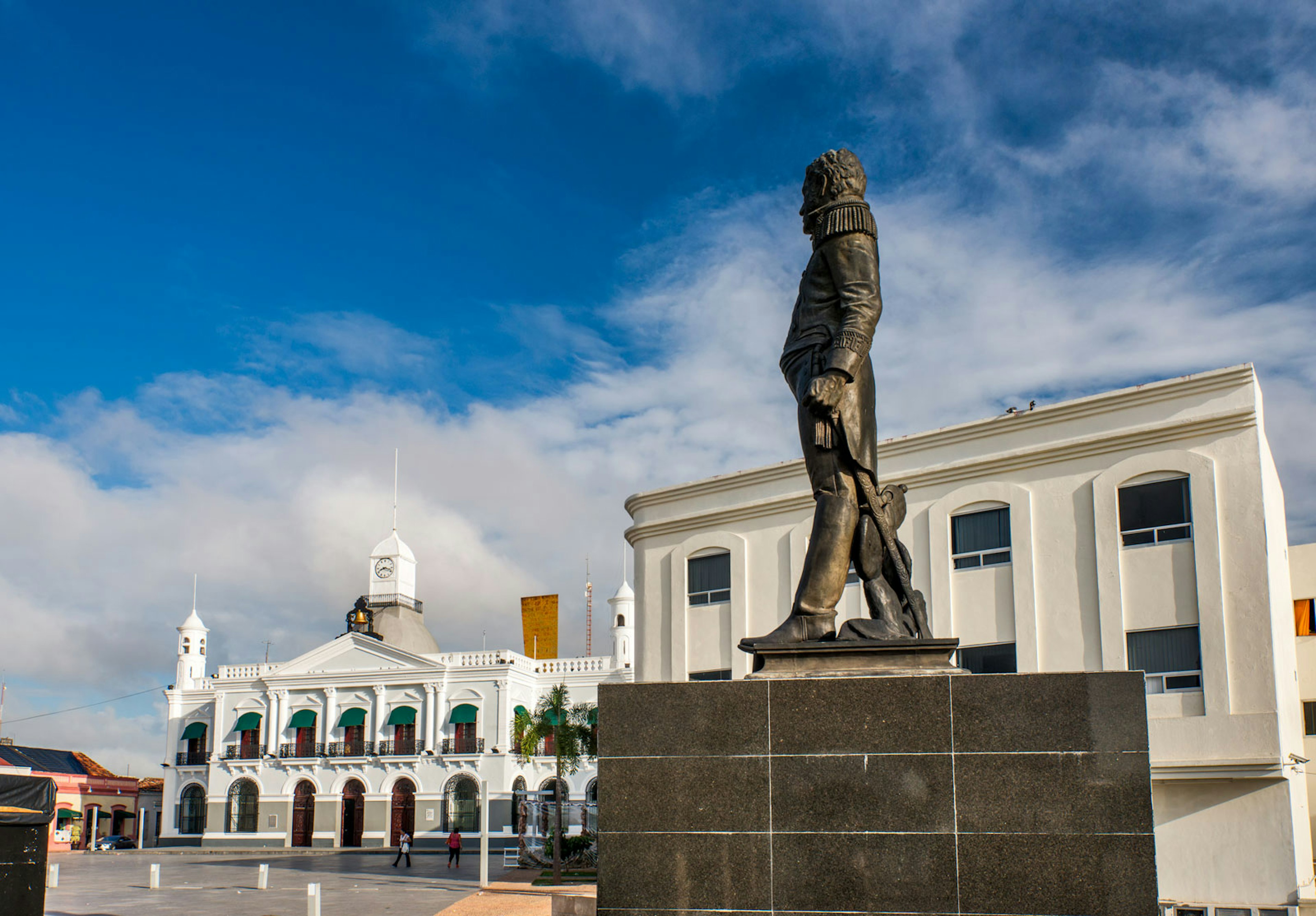 Bronze statue of a man in profile stands before white colonial buildings