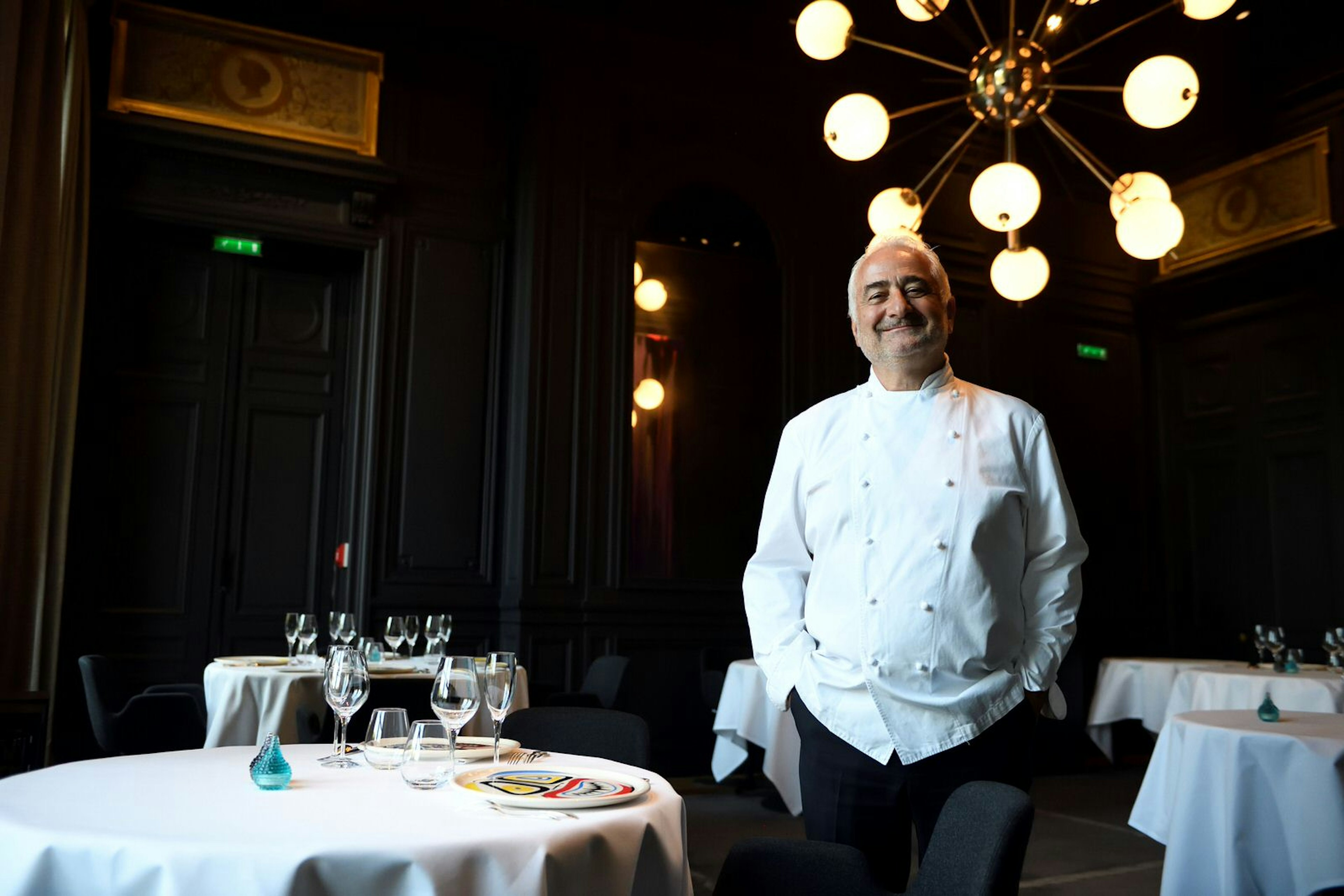 French chef Guy Savoy smiles at the camera wearing his chef's jacket in the dining room of his Michelin three-starred restaurant Guy Savoy de Paris in the Monnaie de Paris. The dark dining room is illuminated by an unusual, globular light fixture.