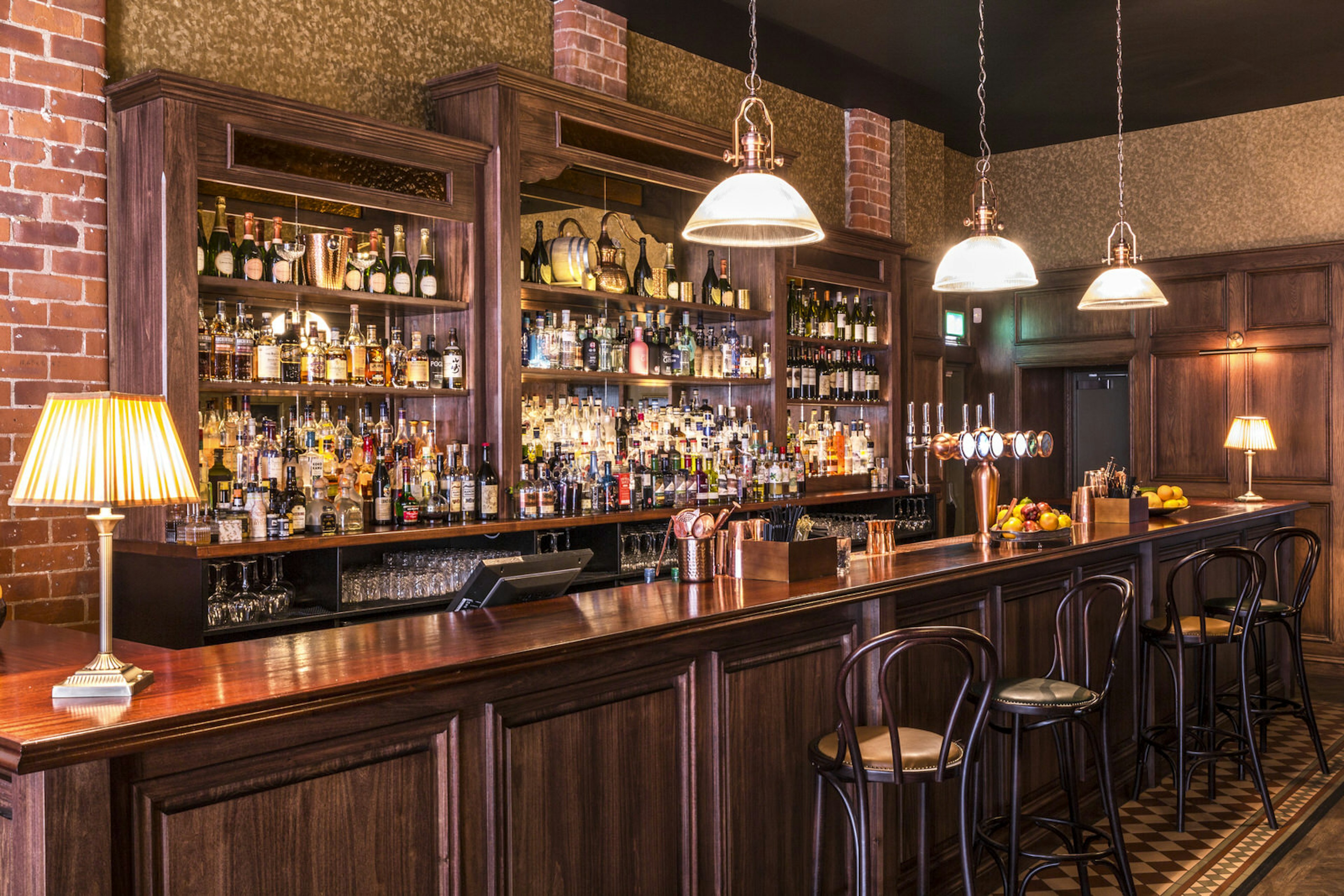 A dark wood bar with rows and rows of spirit bottles in the background, and three lights hanging low over the bar