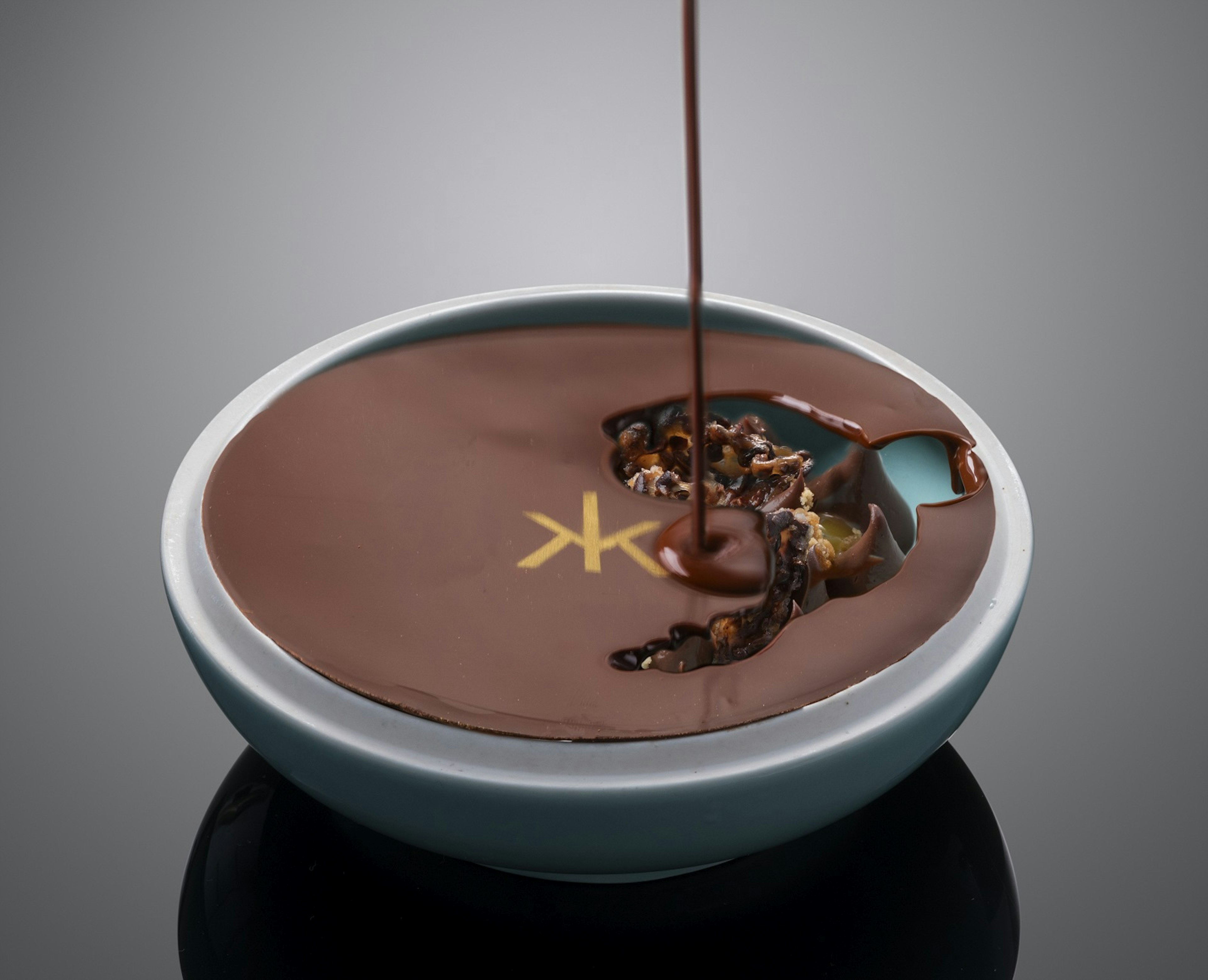 An indulgent Slovenian chocolate pot, featuring the Hakkasan logo in gold. The hard chocolate surface is being drizzled with a hot chocolate sauce to melt the top and reveal the insides.