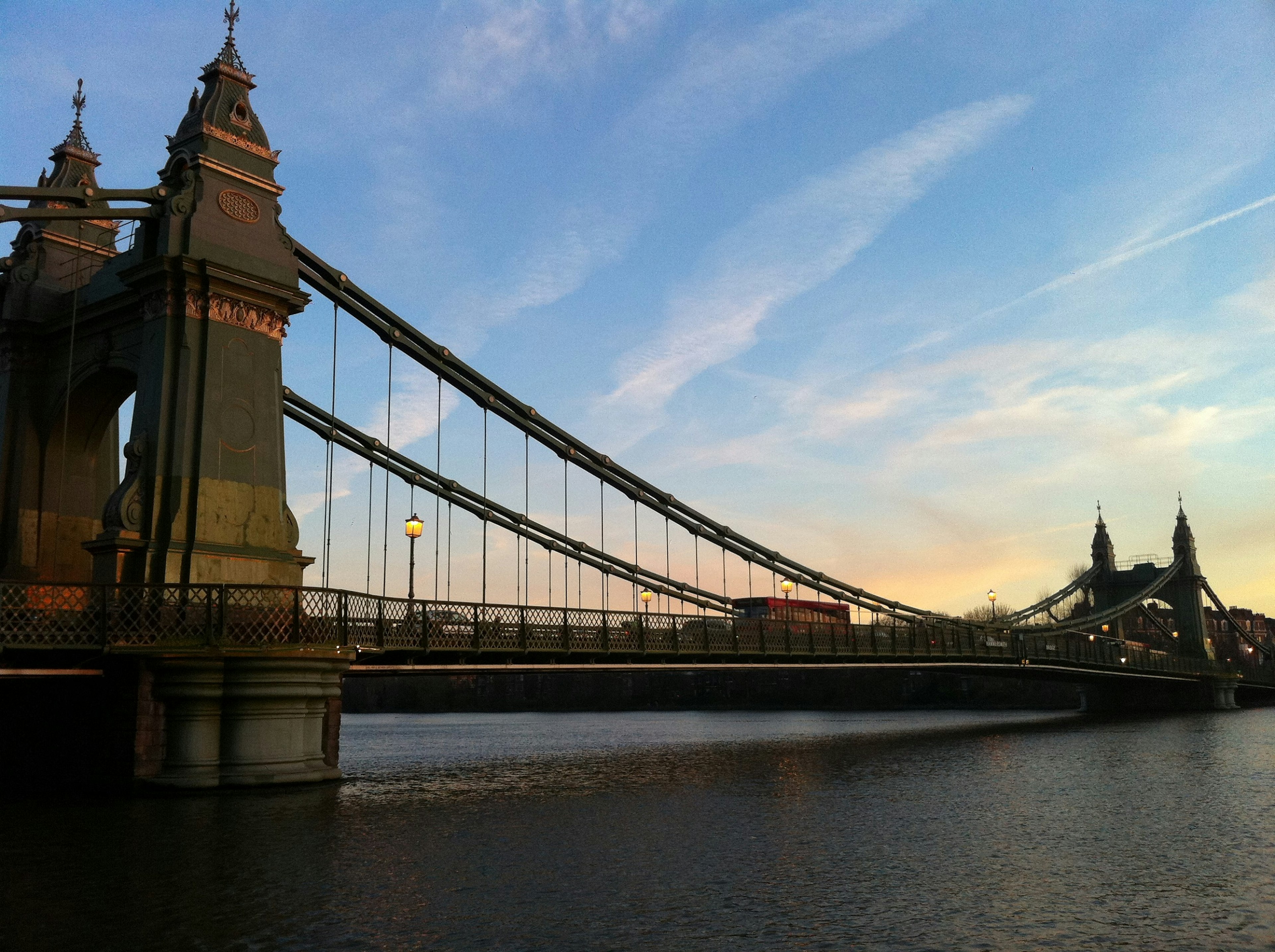 An ornate iron bridge suspension bridge spans the River Thames; the structure of the suspension elements look like massive bicycle chains