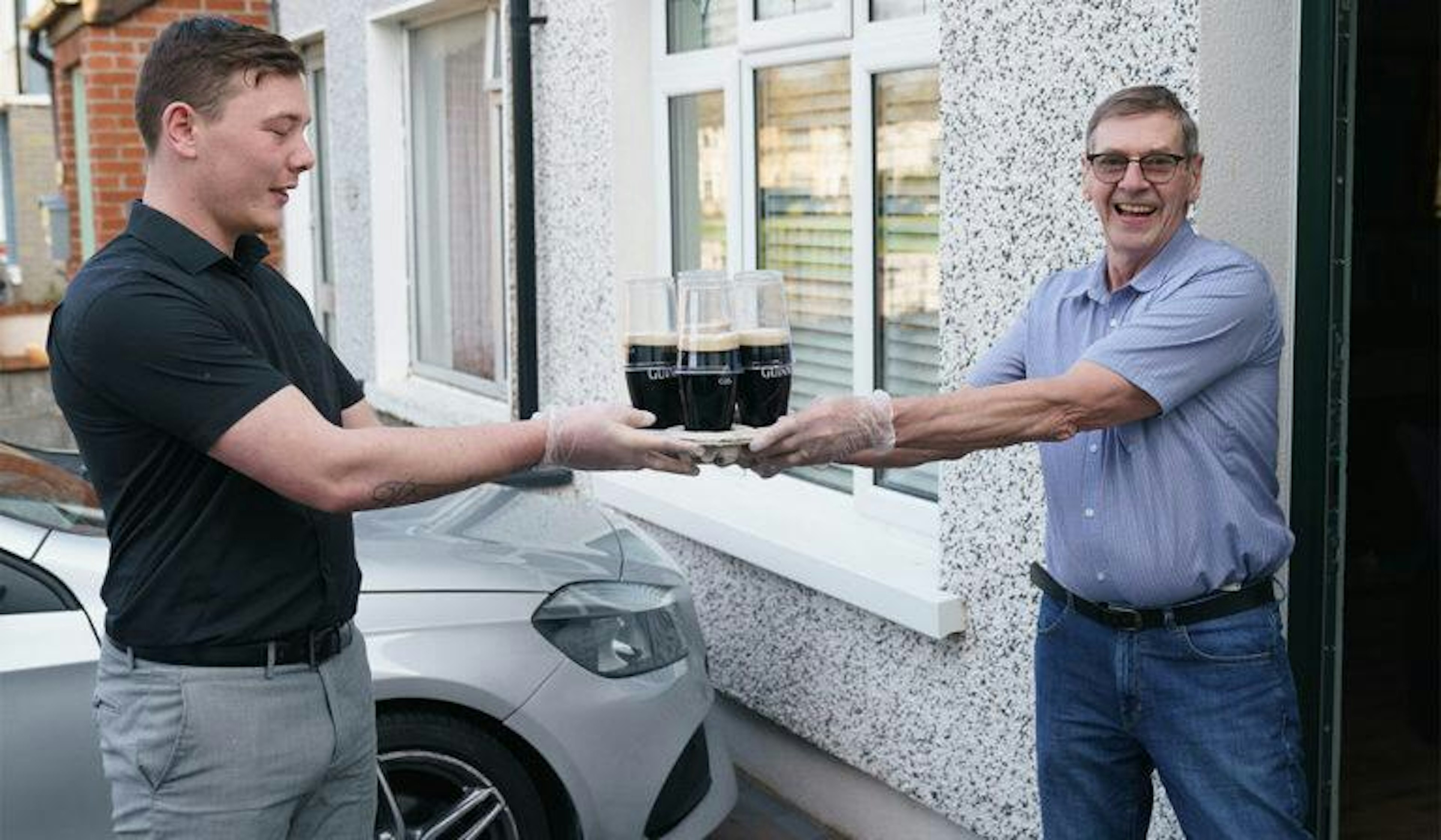 A young man hands over a tray of pints to an older man. Both are smiling for the camera.