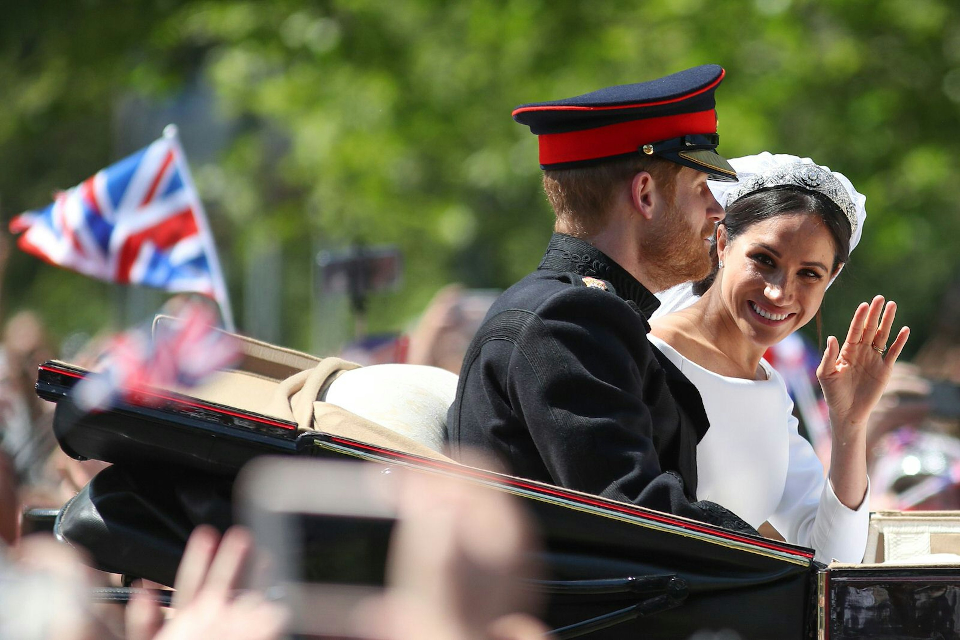 Above a blur of waving hands from gathered crowds is the open-topped carriage carrying Prince Harry and Meghan Markle; Harry in uniform looks forward while Meghan has her head turned towards to crowd and camera, with a bright smile and wave; a blurred British flag flies in the background against a forested backdrop © DANIEL LEAL-OLIVAS / Contributor / Getty Images