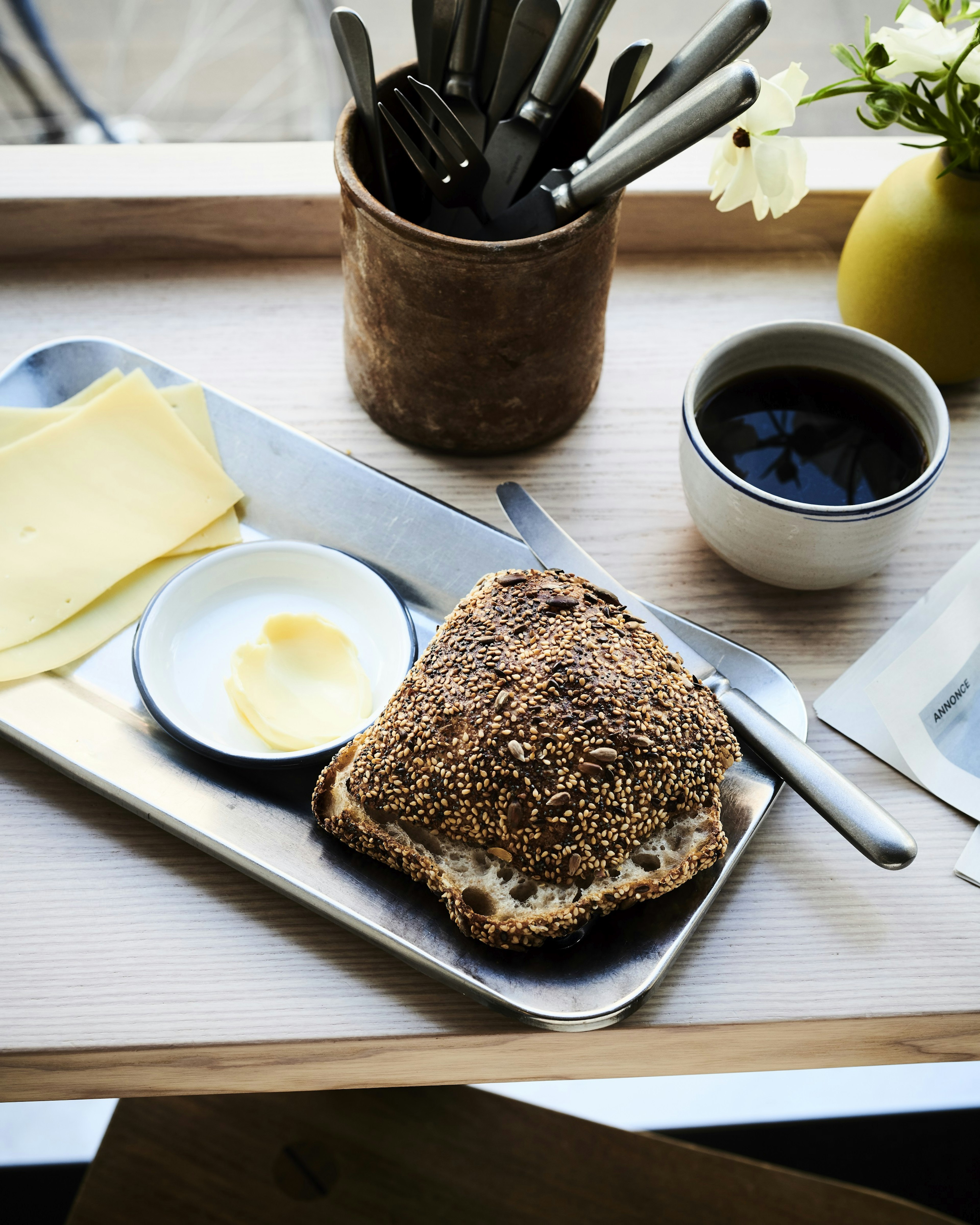 A silver tray sits on a wooden bar shelf; on the plate is some fresh bread, next to slices of cheese and a tiny bowl with butter.