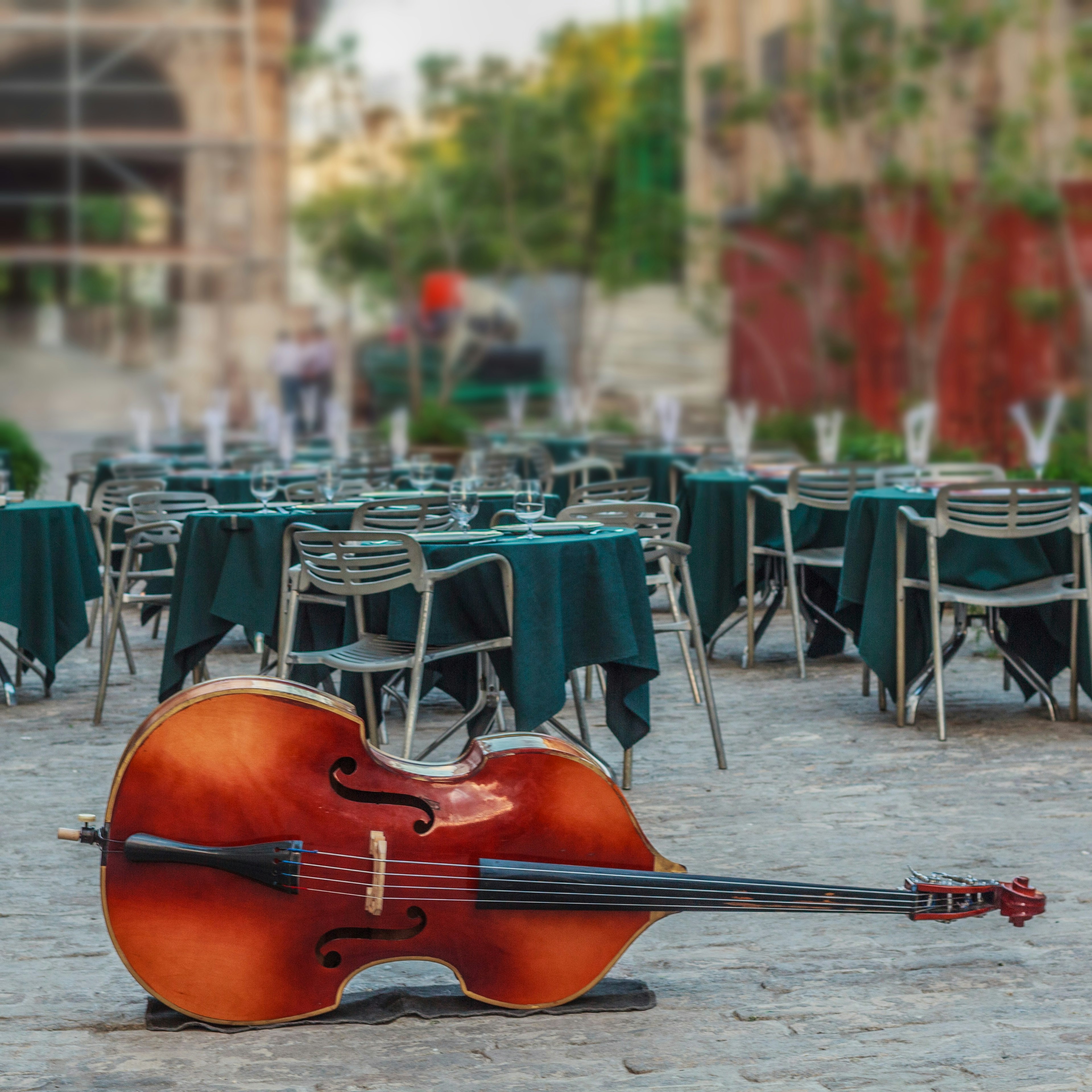 A large cello bass lies on its side in front of a group of tables positioned throughout a open-air cafe; Havana anniversary