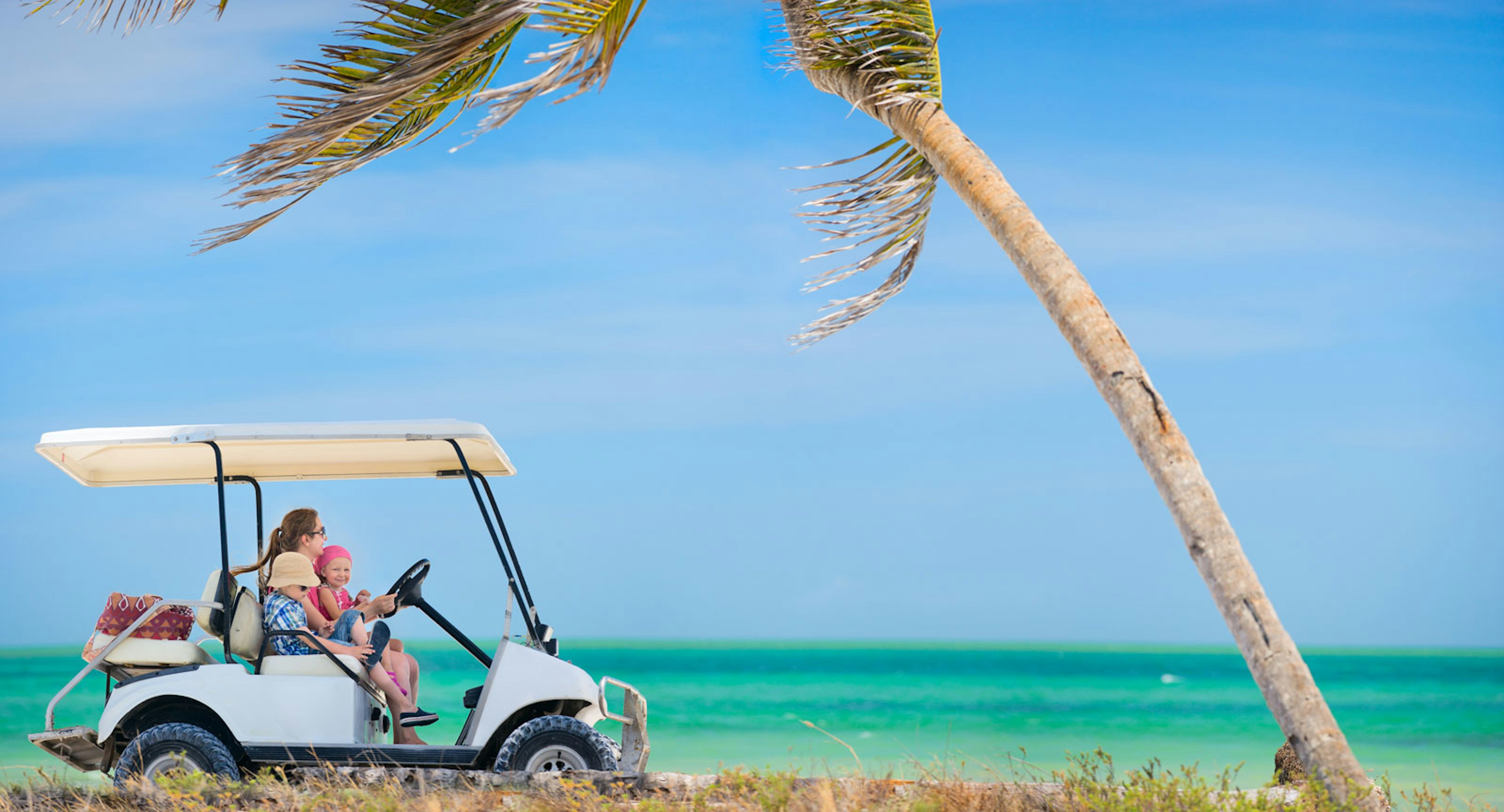 a woman drives a golf cart with a young girl on her lap and a boy next to her on the beach toward a palm tree