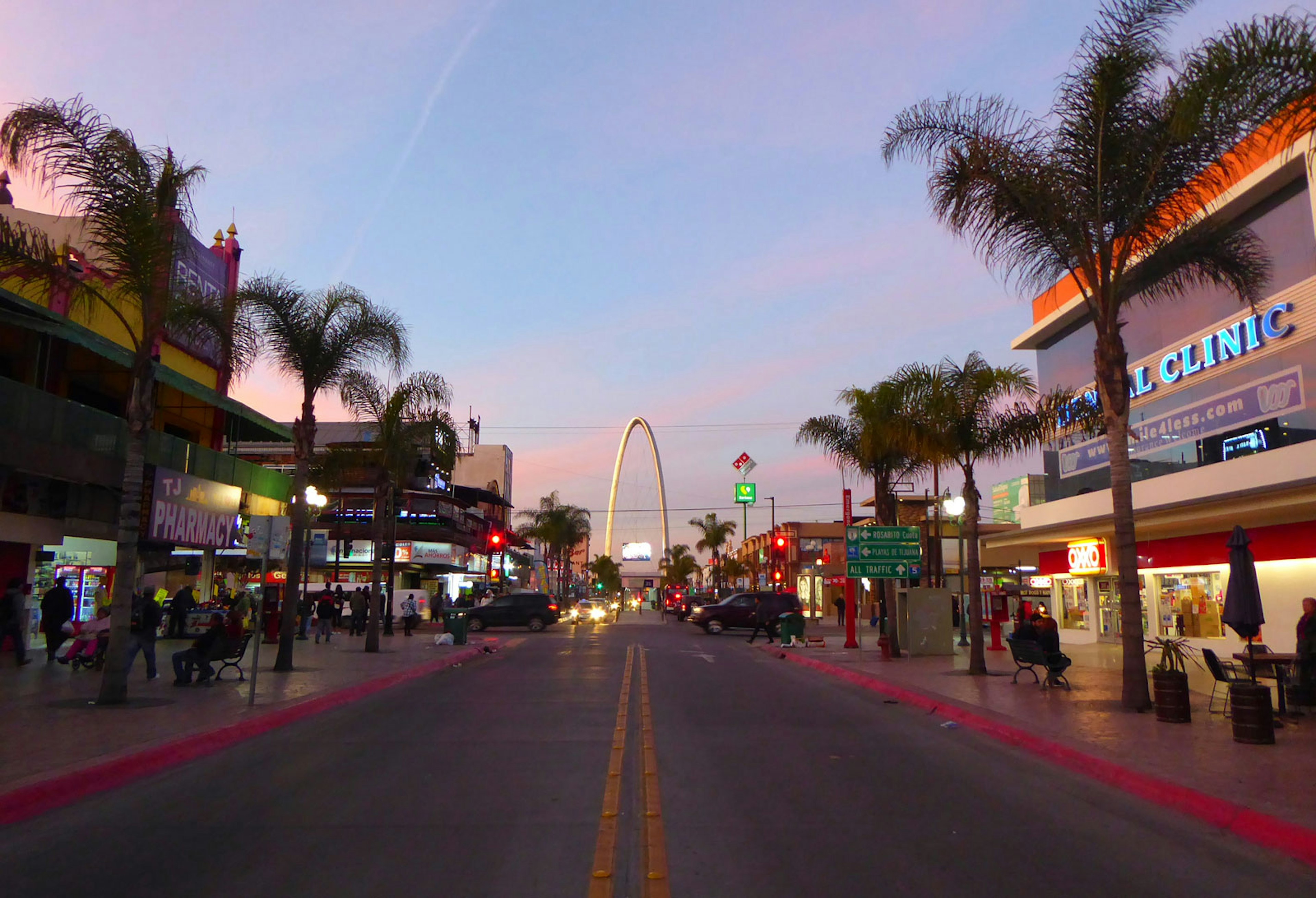 An empty street lined with palm trees with an arch at one end under a blue sky dotted with pink clouds