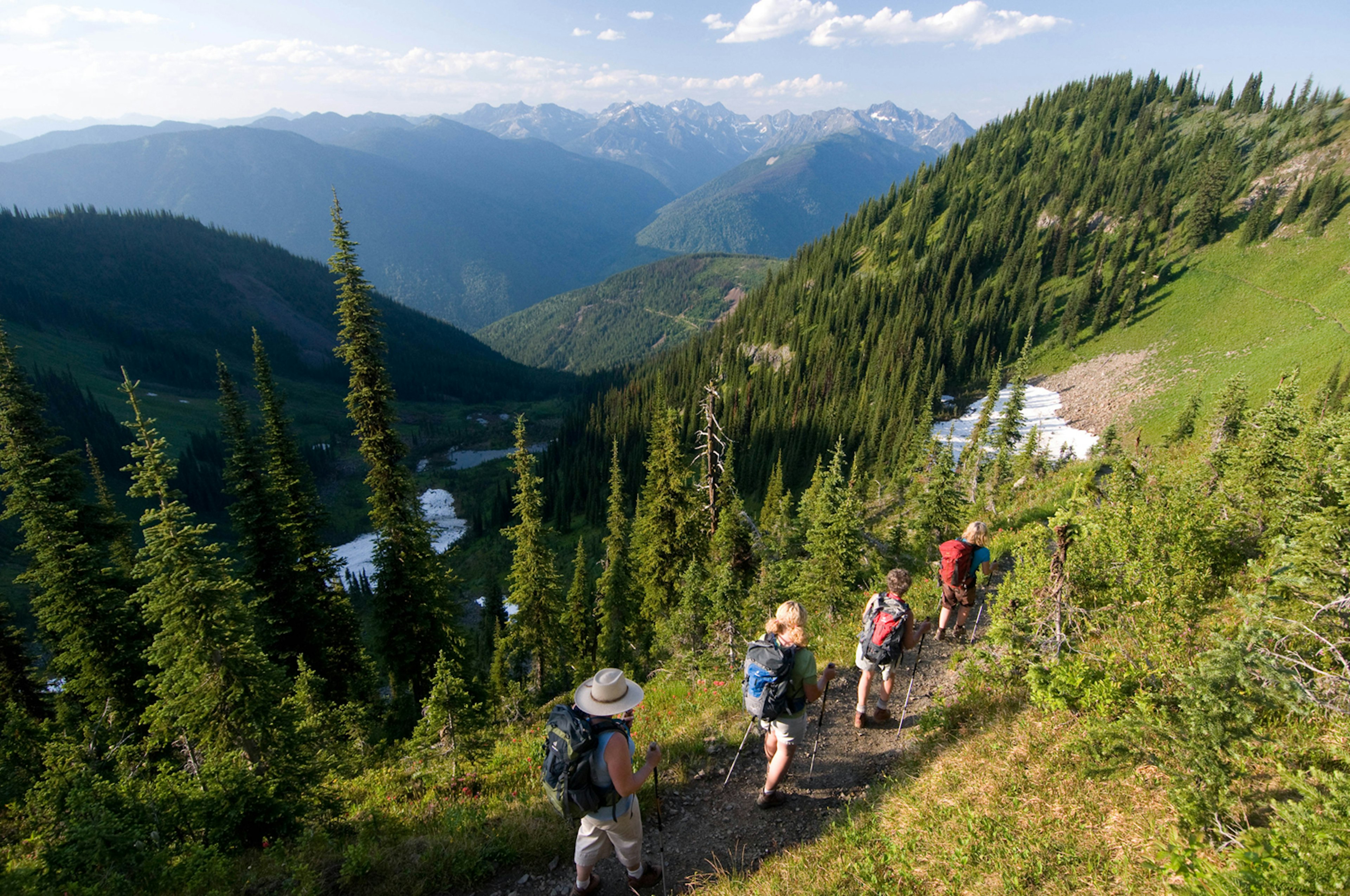 Four people are hiking on a forested mountain trail. It's a bright day but there are some patches of snow on the ground. Beyond the group is a rugged mountain range.