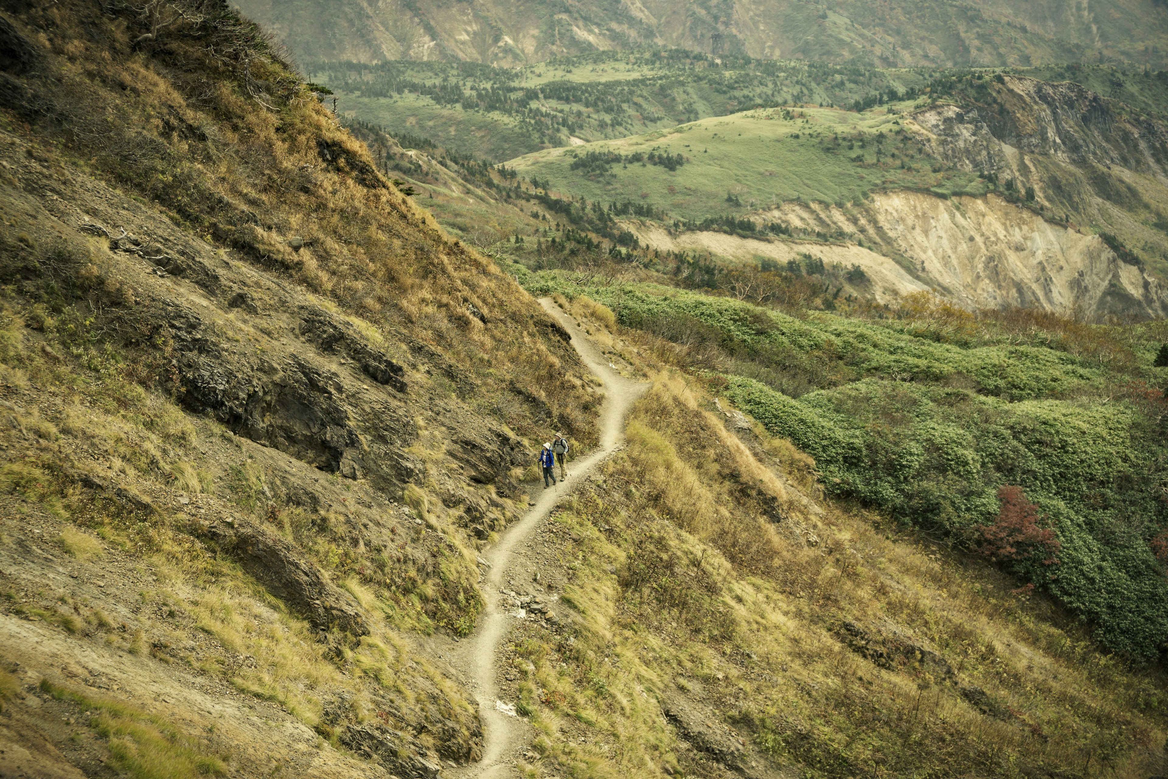 Two hikers walk along a path running along a hill in the Hakusan National Park. In the background, more forested hills are visible.