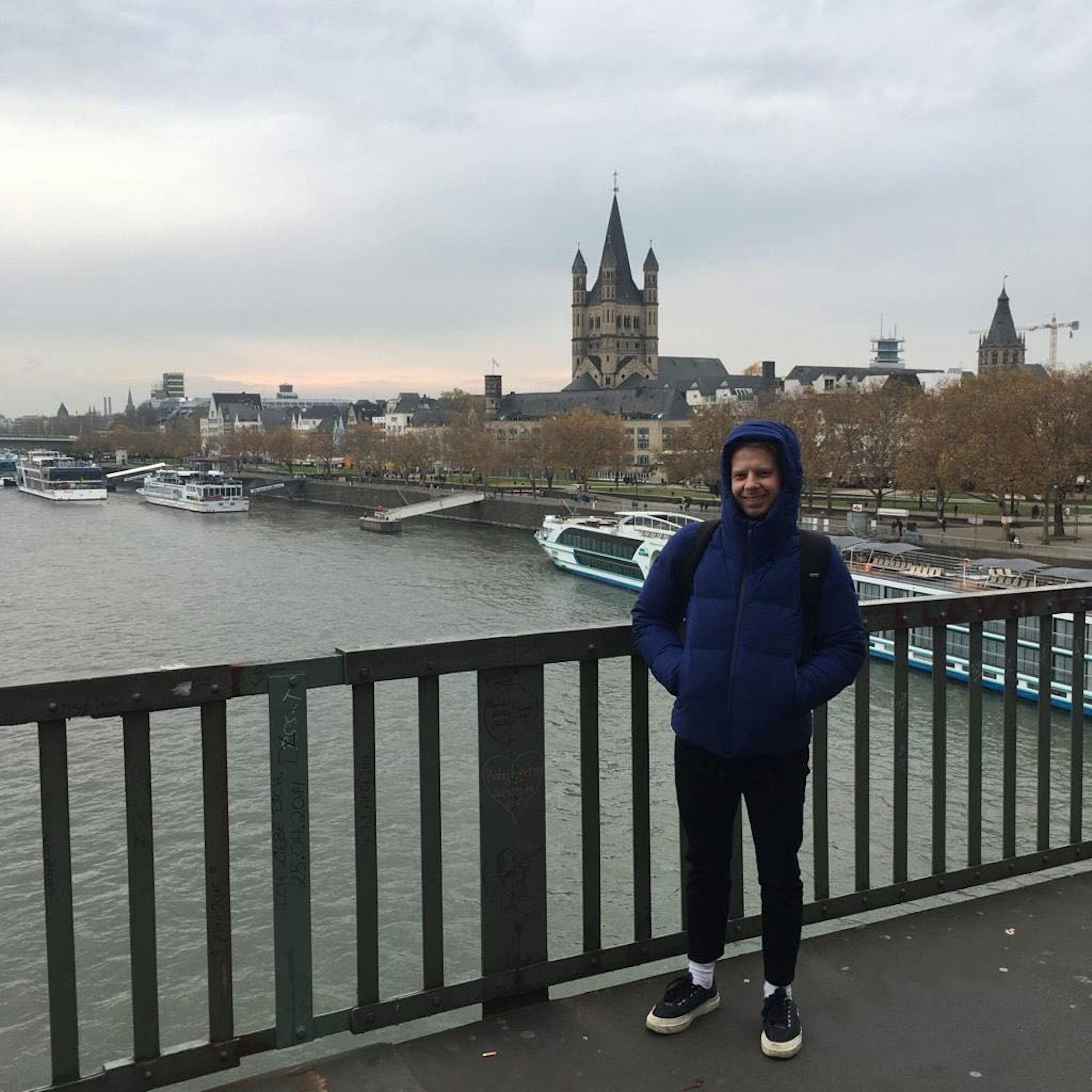 The author stands on a bridge wearing a thick blue jacket with his hood up. In the background is a river with canal boats on it.