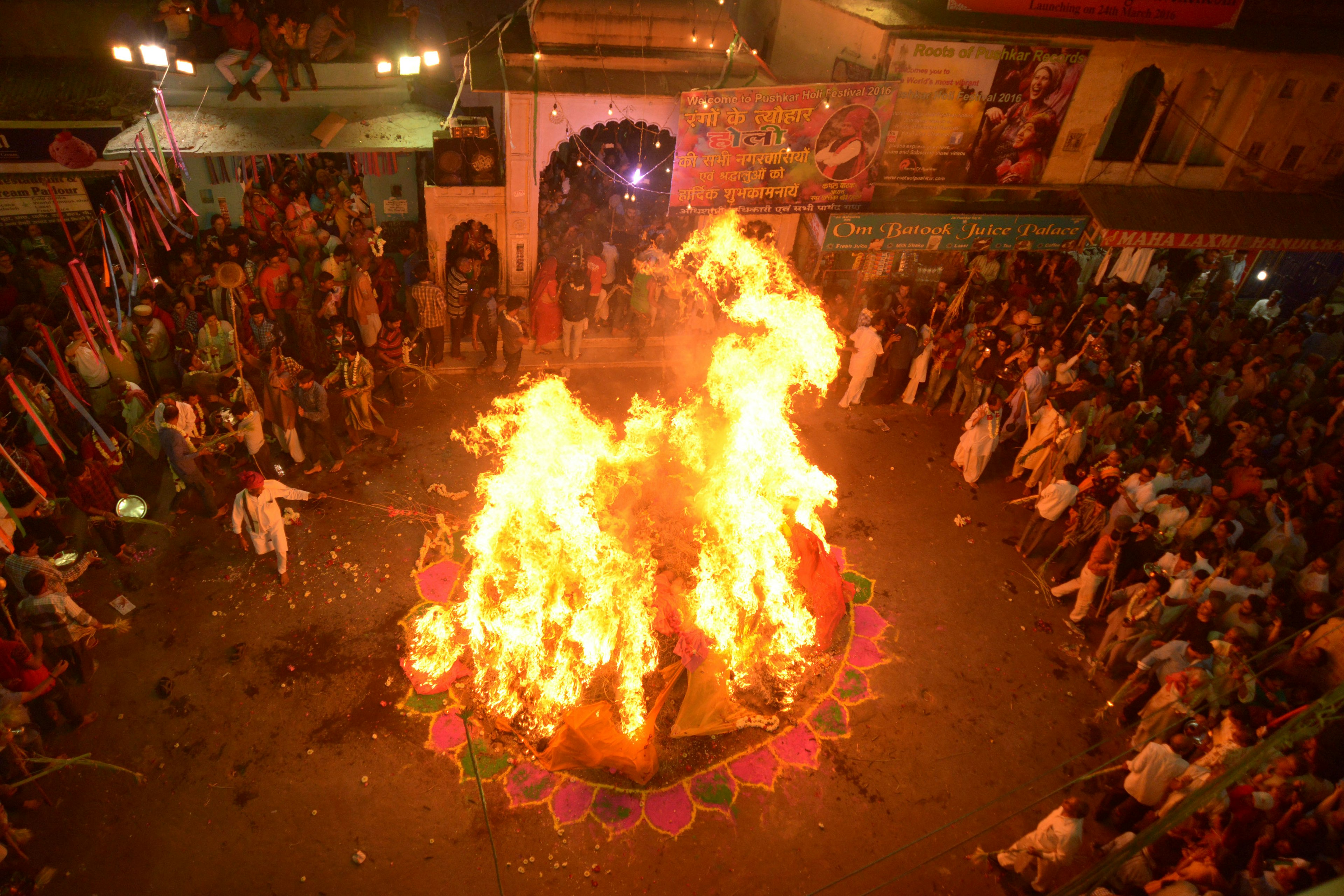 A giant bonfire rages in the middle of a street in Pushkar, India, with crowds circled around it. A man in all white stokes the fire with a long stick.