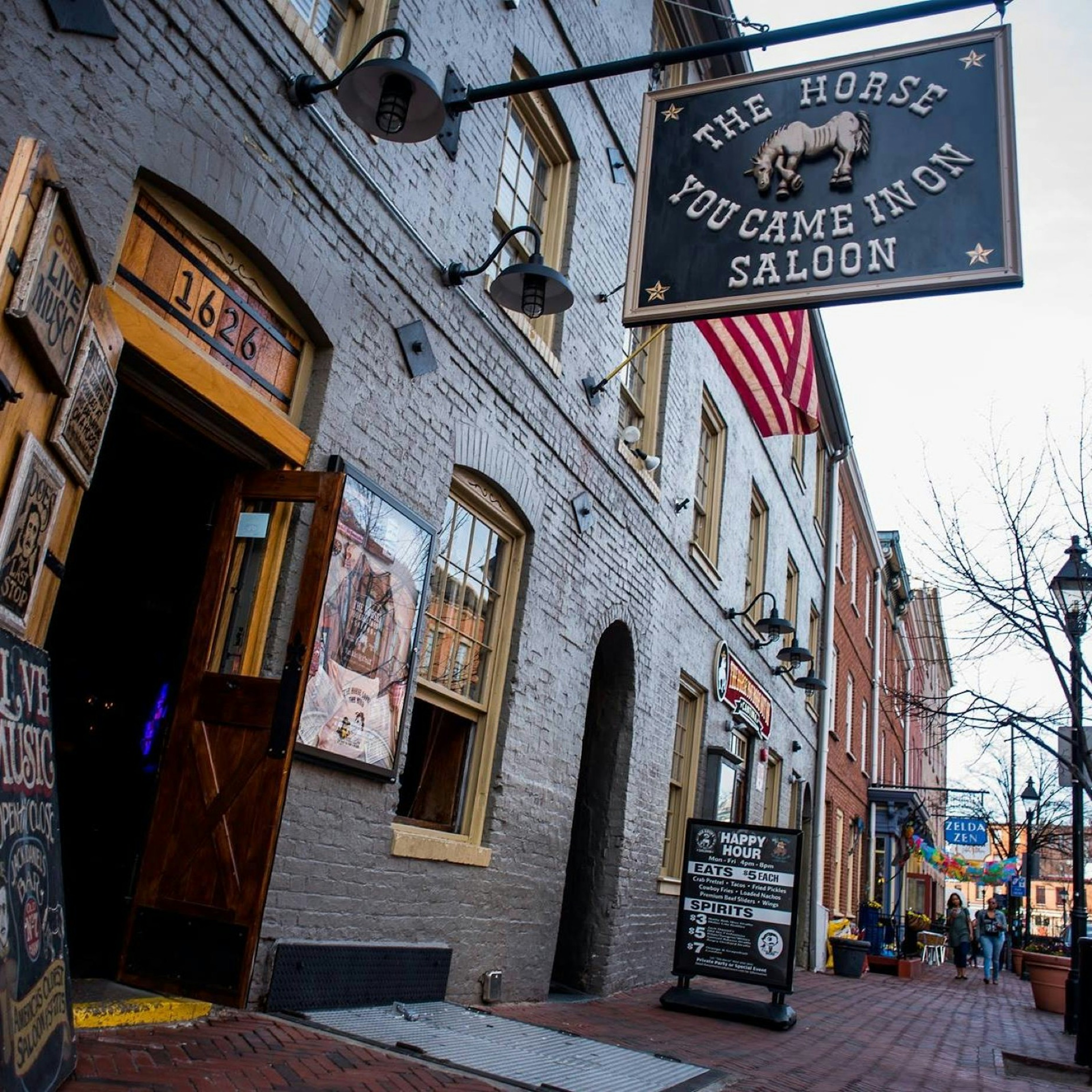 A Georgian brick building is the home of a saloon with a large black sign hanging over the brick street