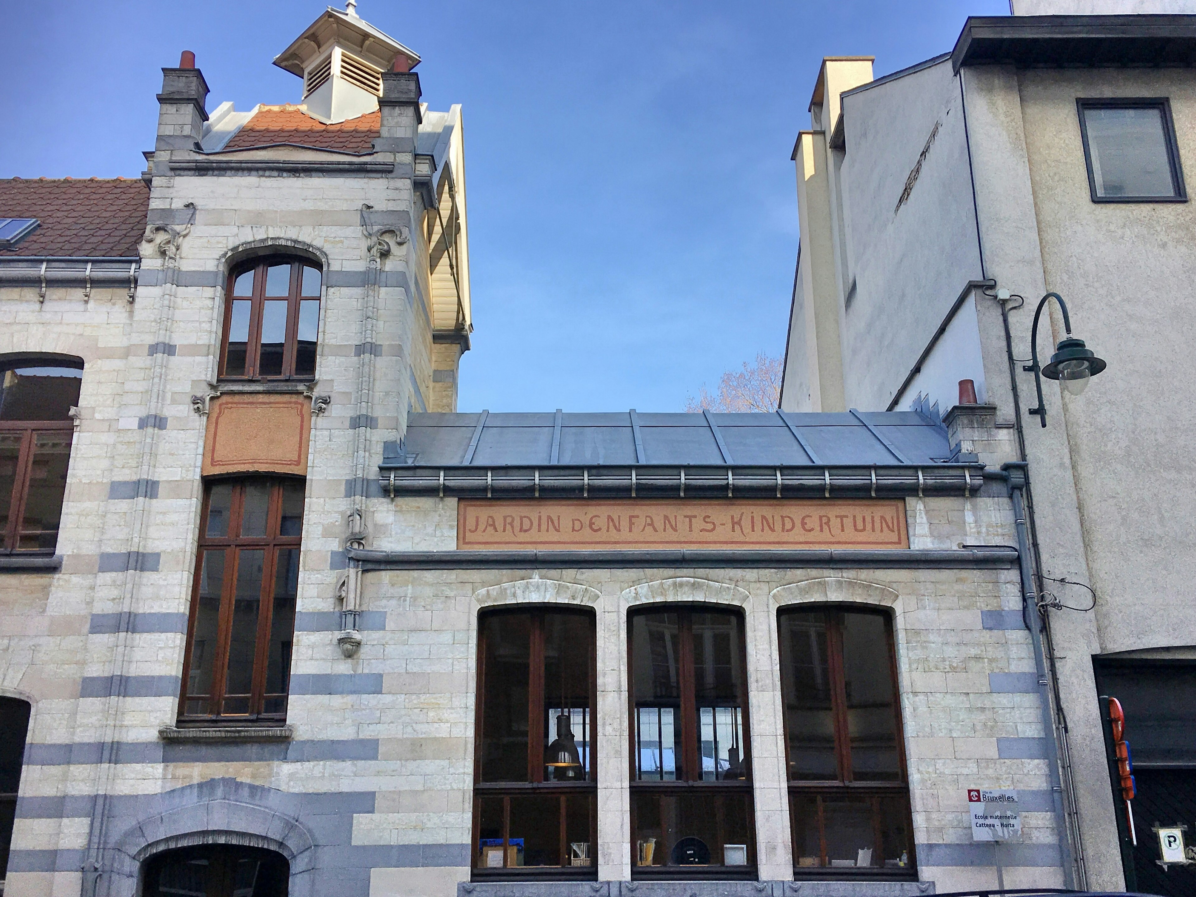 A stone Art Nouveau building, with bricks in varying shades of grey, and an orange sign saying Jardin d’Enfants-Kindertuin; there is a grey roof, leaded windows and chimneys, and it adjoins similar buildings.