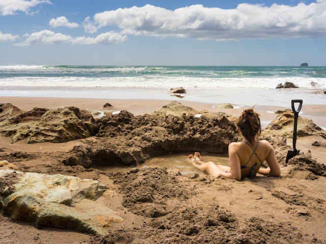 A woman sitting in a self-dug hot pool on a sandy beach looking out to sea