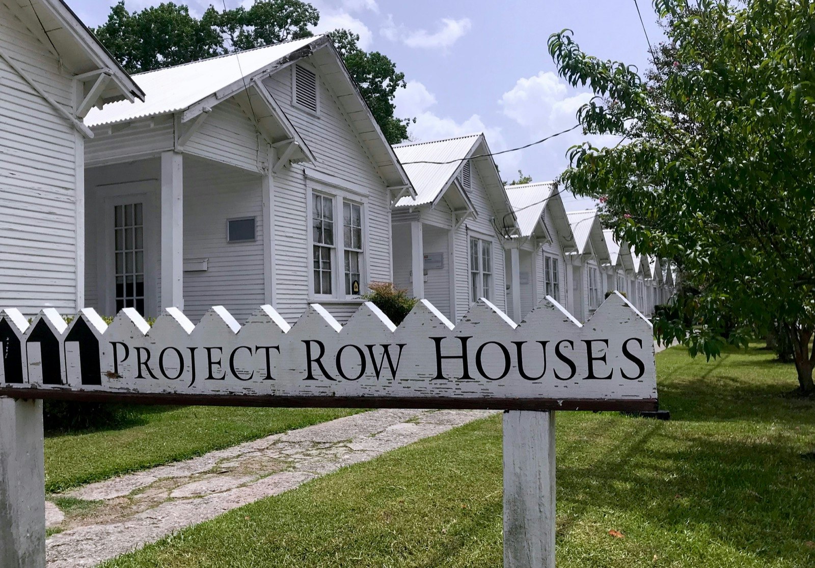 A line of identical small white-painted houses, with a sidewalk in front, is framed by a sign that reads 'Project Row Houses' © Lisa Dunford / Ĵý