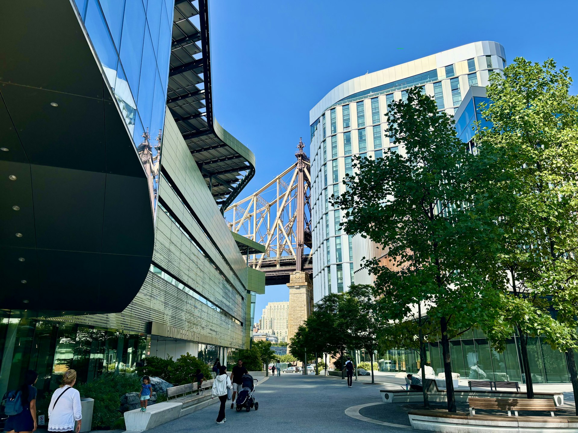 People walk path through the high-tech university buildings of the Cornell Tech campus, with a view of the Queensboro Bridge in the distance, Roosevelt Island, New York City