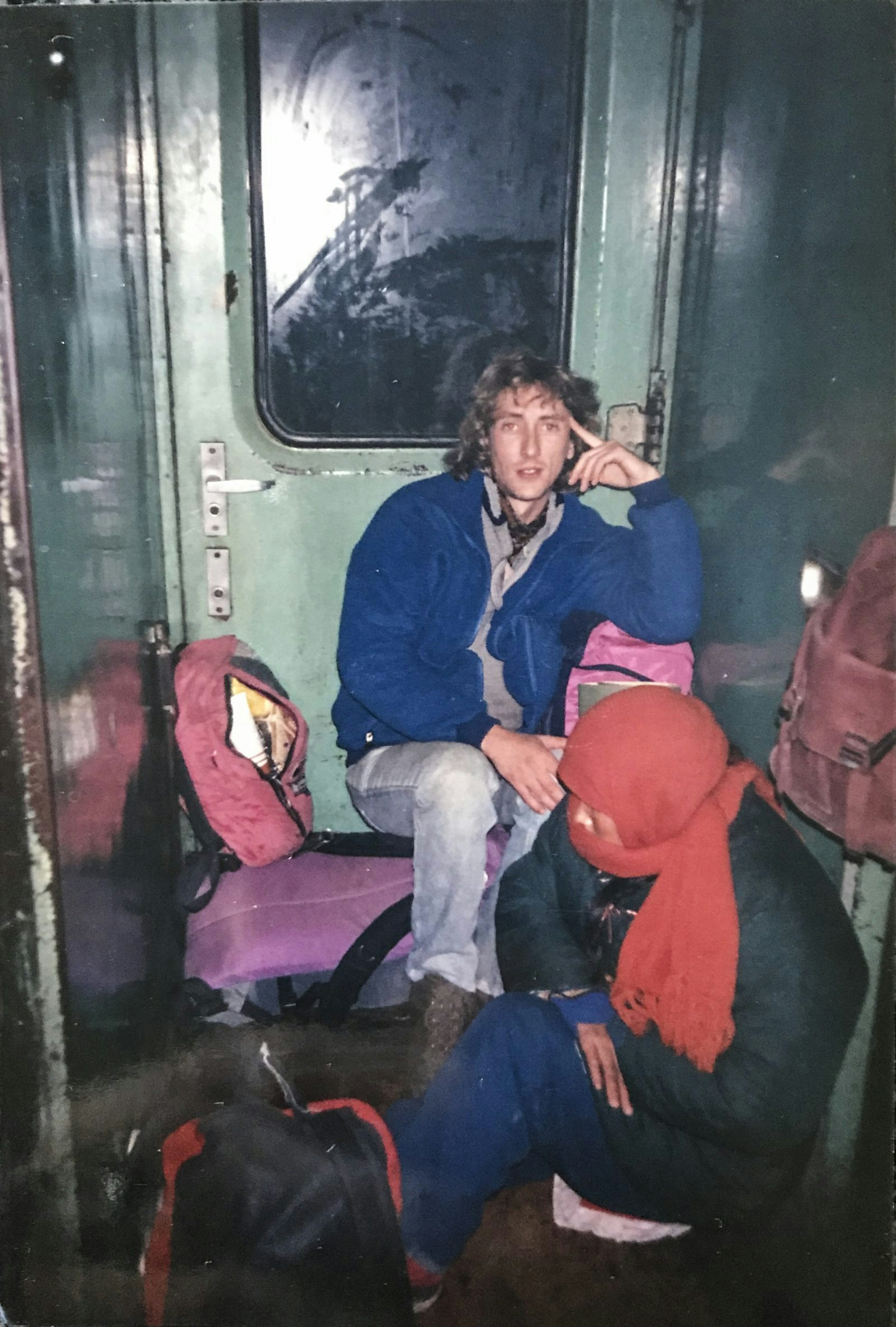 Steve and another person sit in a narrow part of a train by a doorway. They have bags around them and look uncomfortable. Ice is visible on the edge of the door.