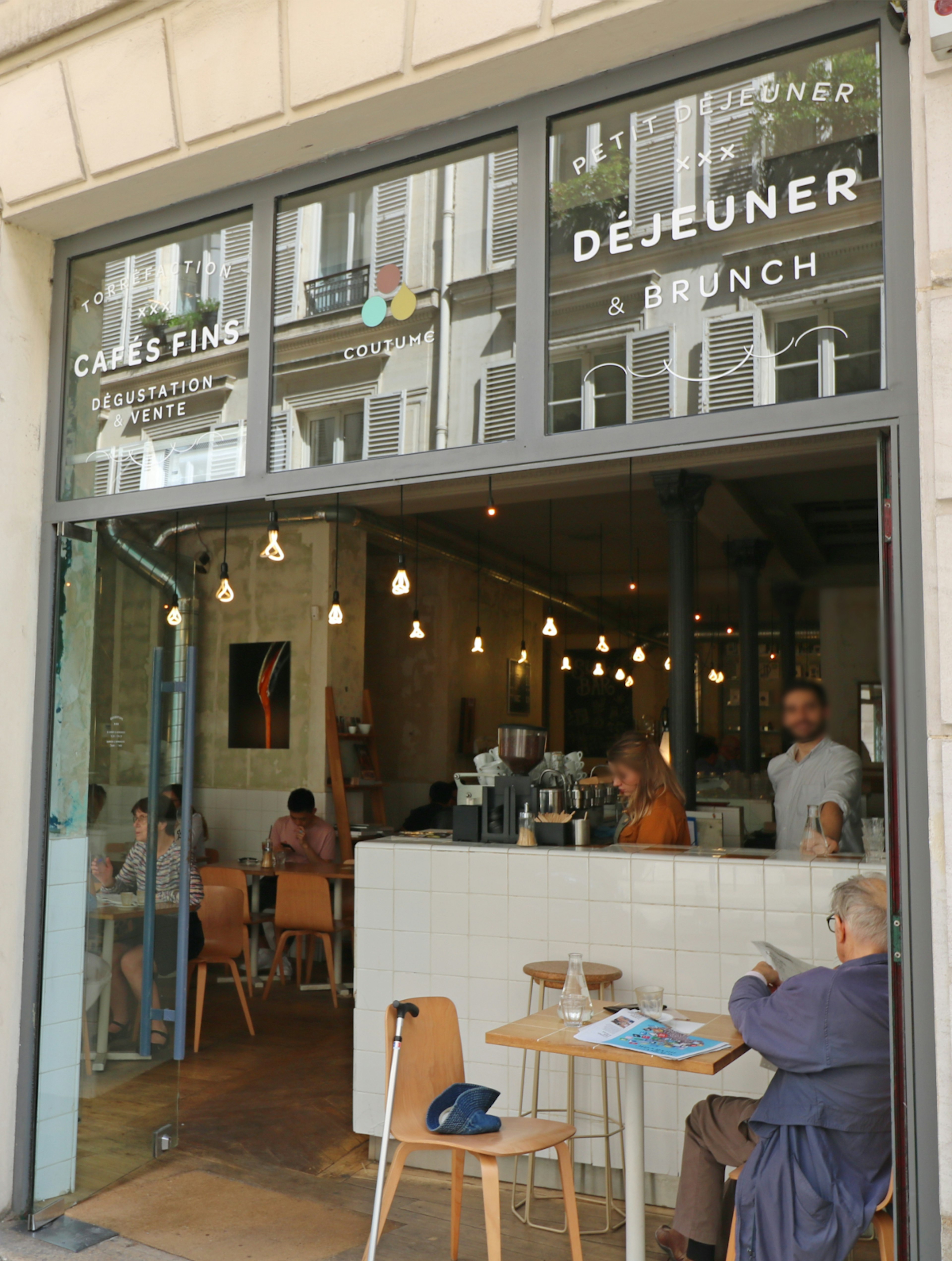 A man sits on a table outside of Coutume Café in Paris; the cafe has a modern glass front, with doors that have been fully opened to reveal a white tiled bar behind which servers are standing.