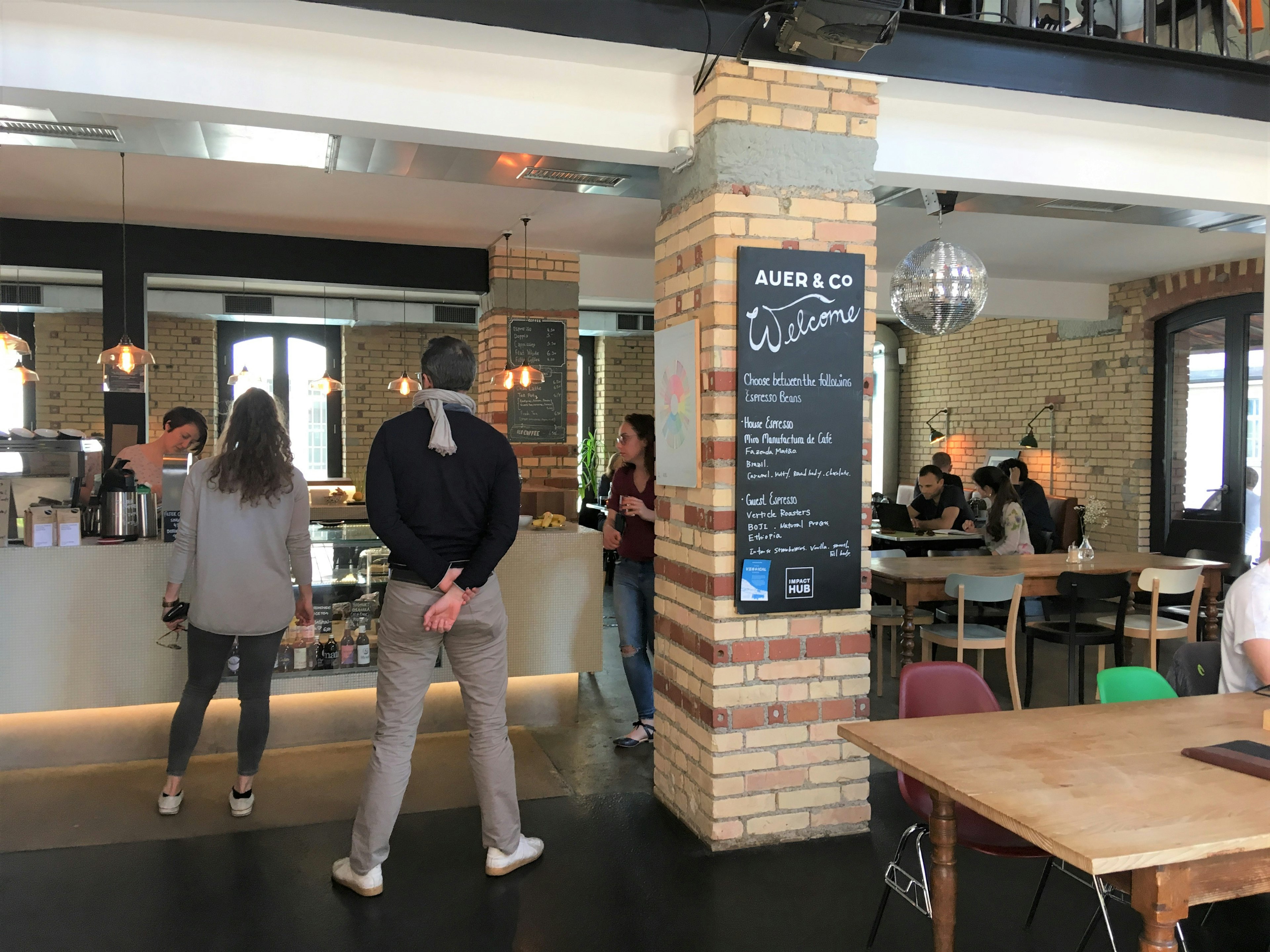 The interior of Auer & Co.; there are exposed brick walls and a brick column in the centre of the room; customers are queuing at the bar while others are working at tables under a glittery silver disco ball.