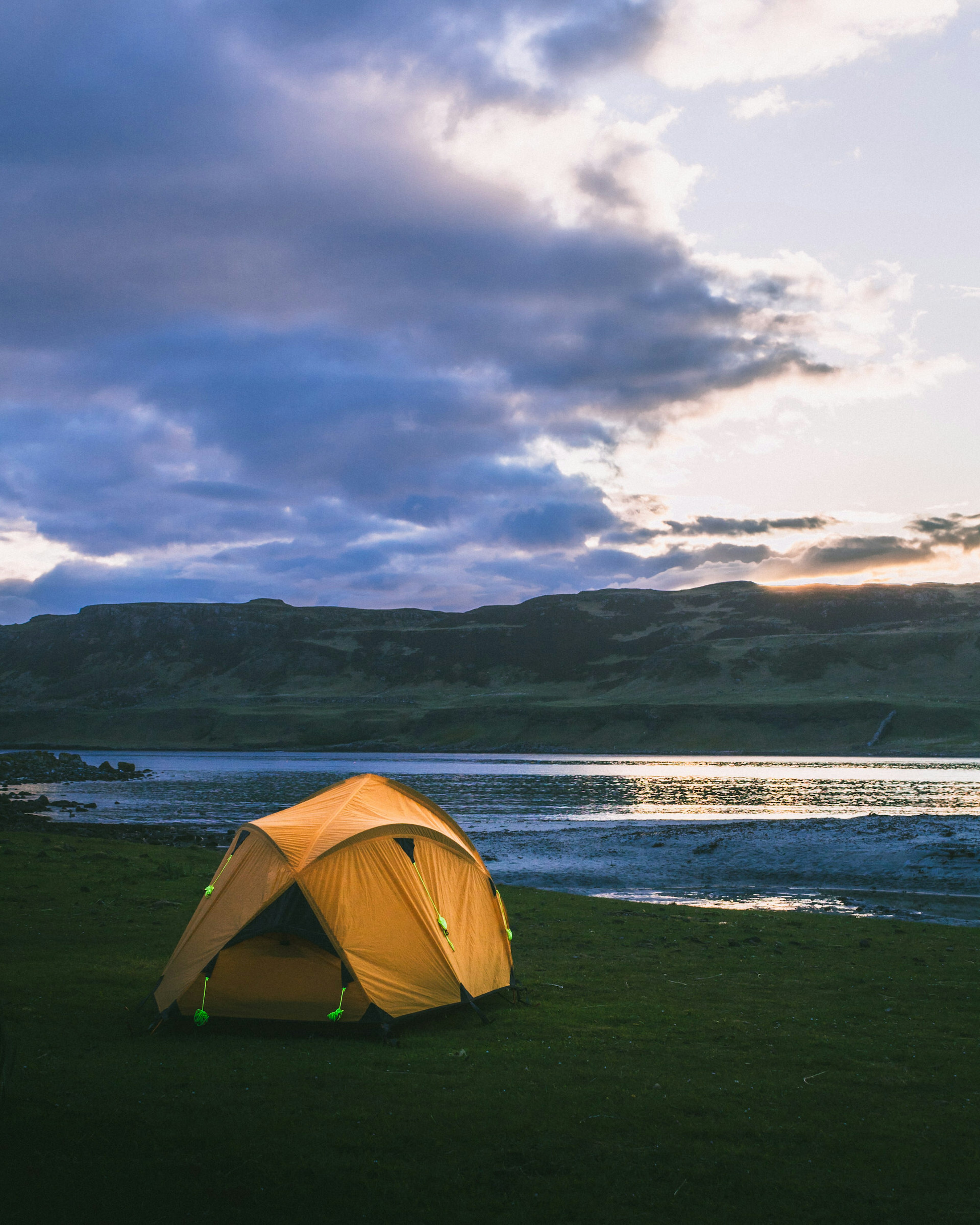 A bright yellow tent has been pitched on grassland just behind a shoreline. It's dusk, and the sun has just dipped below the hills in the distance.