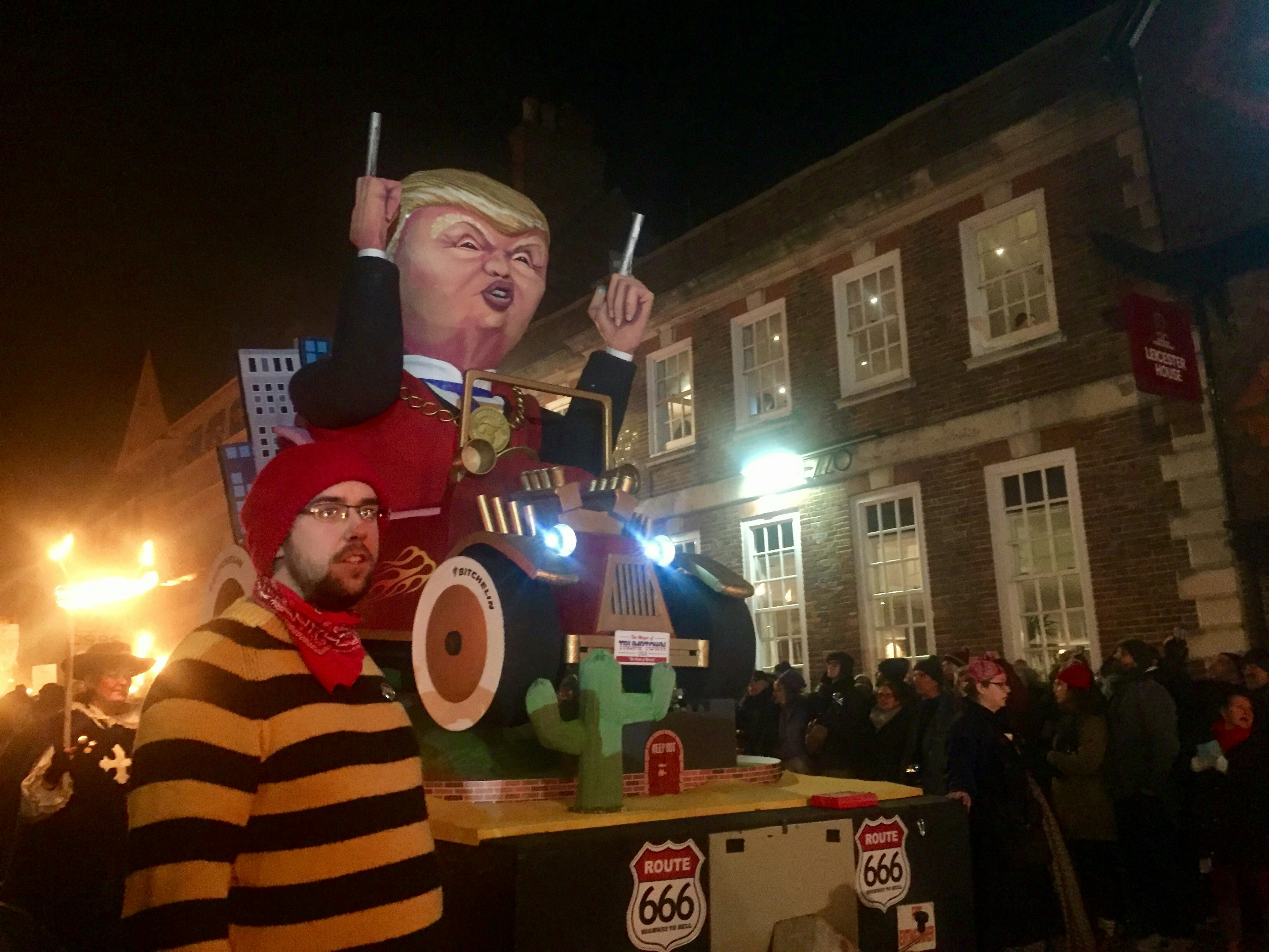 A young man wearing a yellow and black striped t-shirt leads a float with a giant 3D caricature of Donald Trump on it. Trump is depicted toting pistol fingers and is sitting in a car. Crowds are gathered behind.