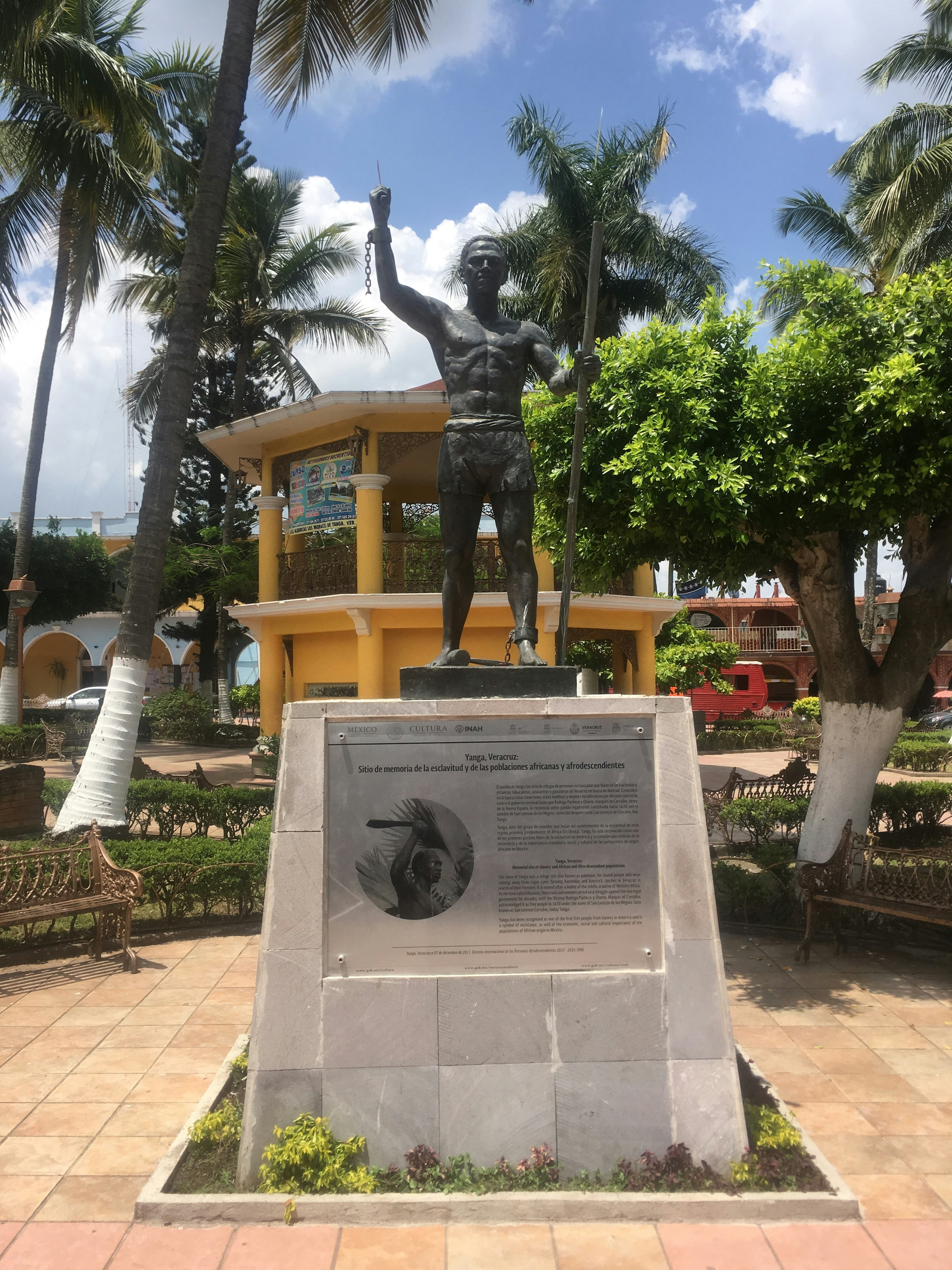 A bronze statue of a shirtless man on a marble plinth with a yellow gazebo in the background surrounded by palm trees