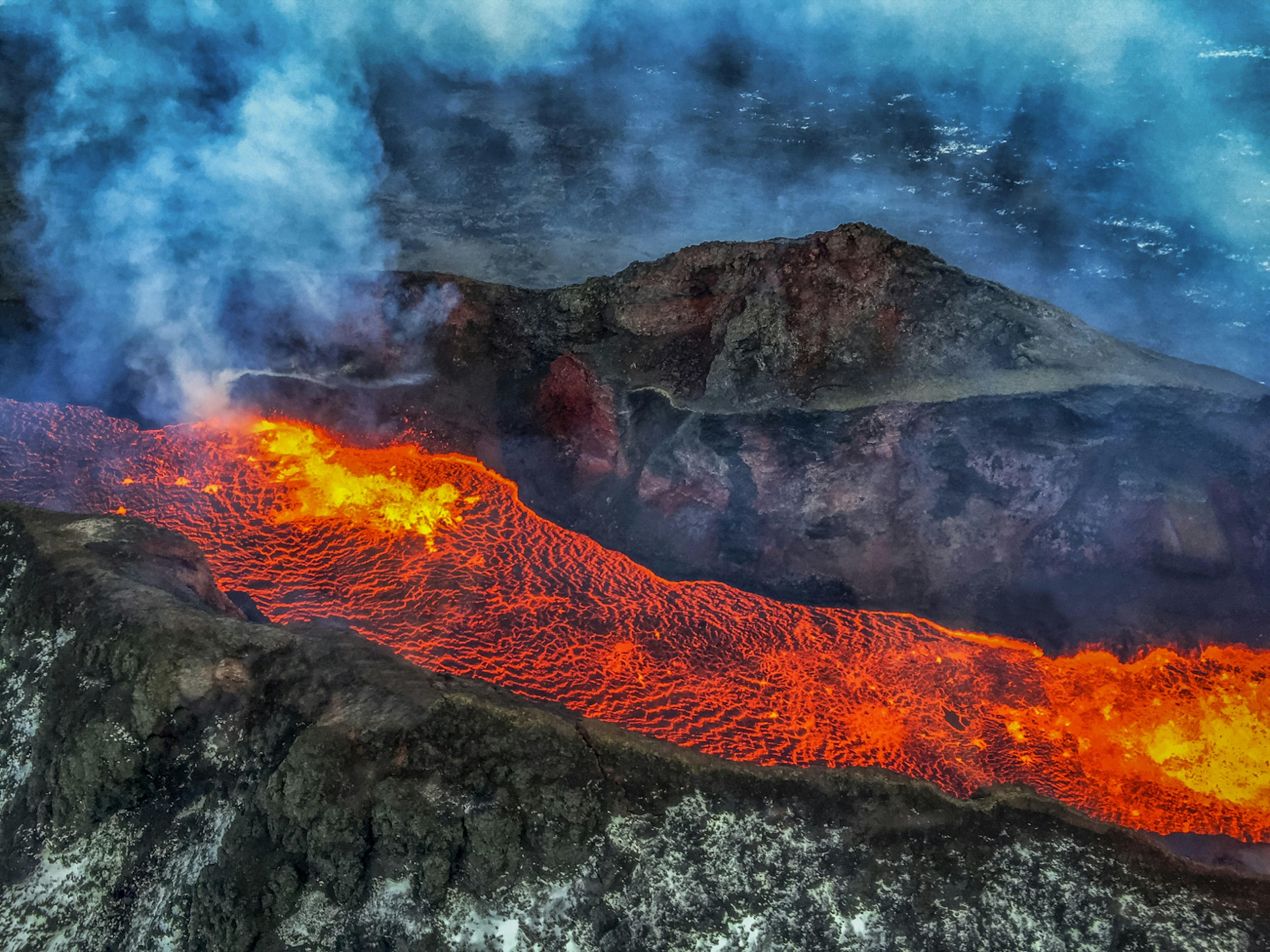 Bright steaming lava flows over dark grey rock, with blue smoke above, in Iceland's interior