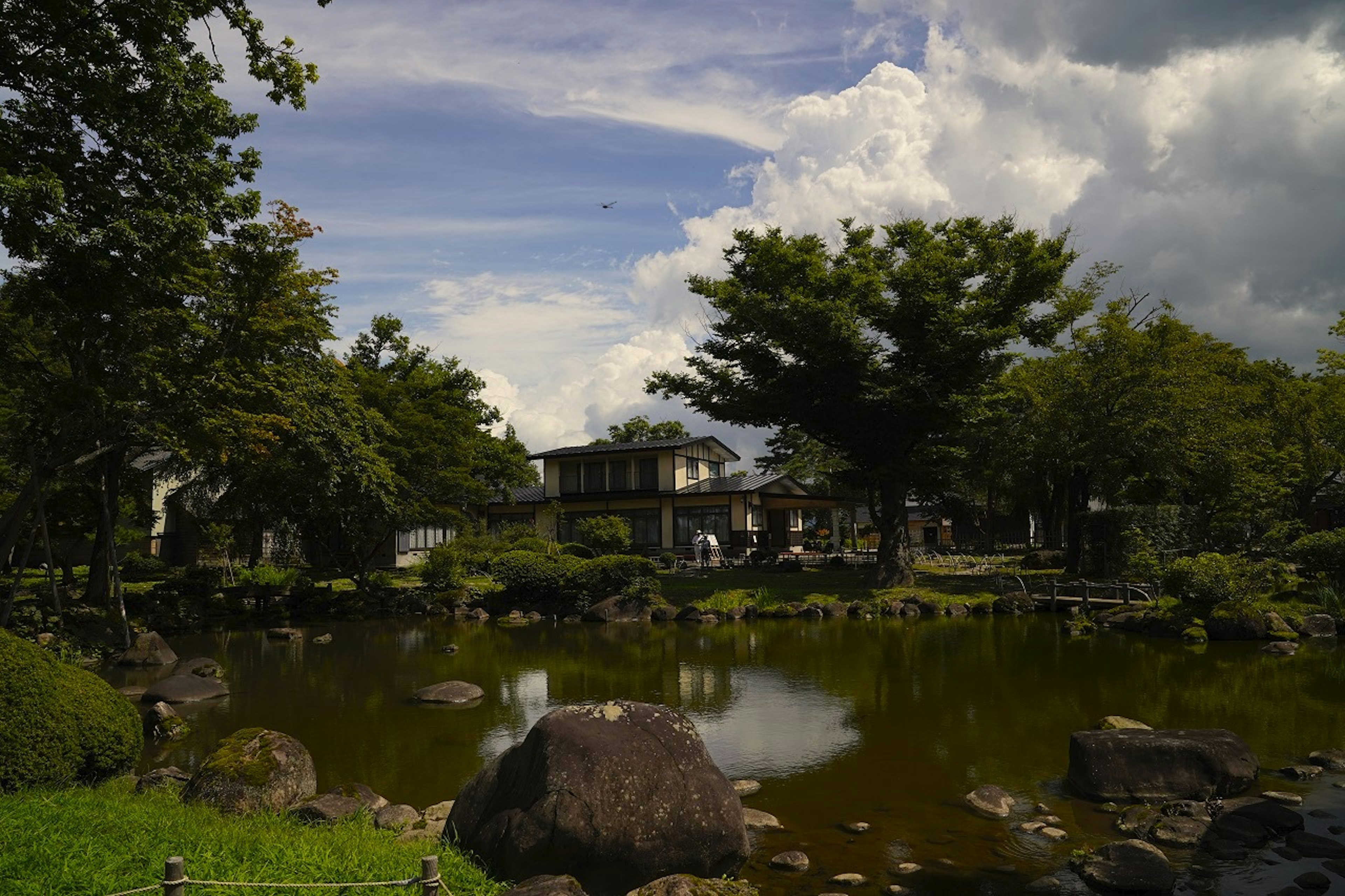 A lake is surrounded by trees and foliage, with a large house in the background