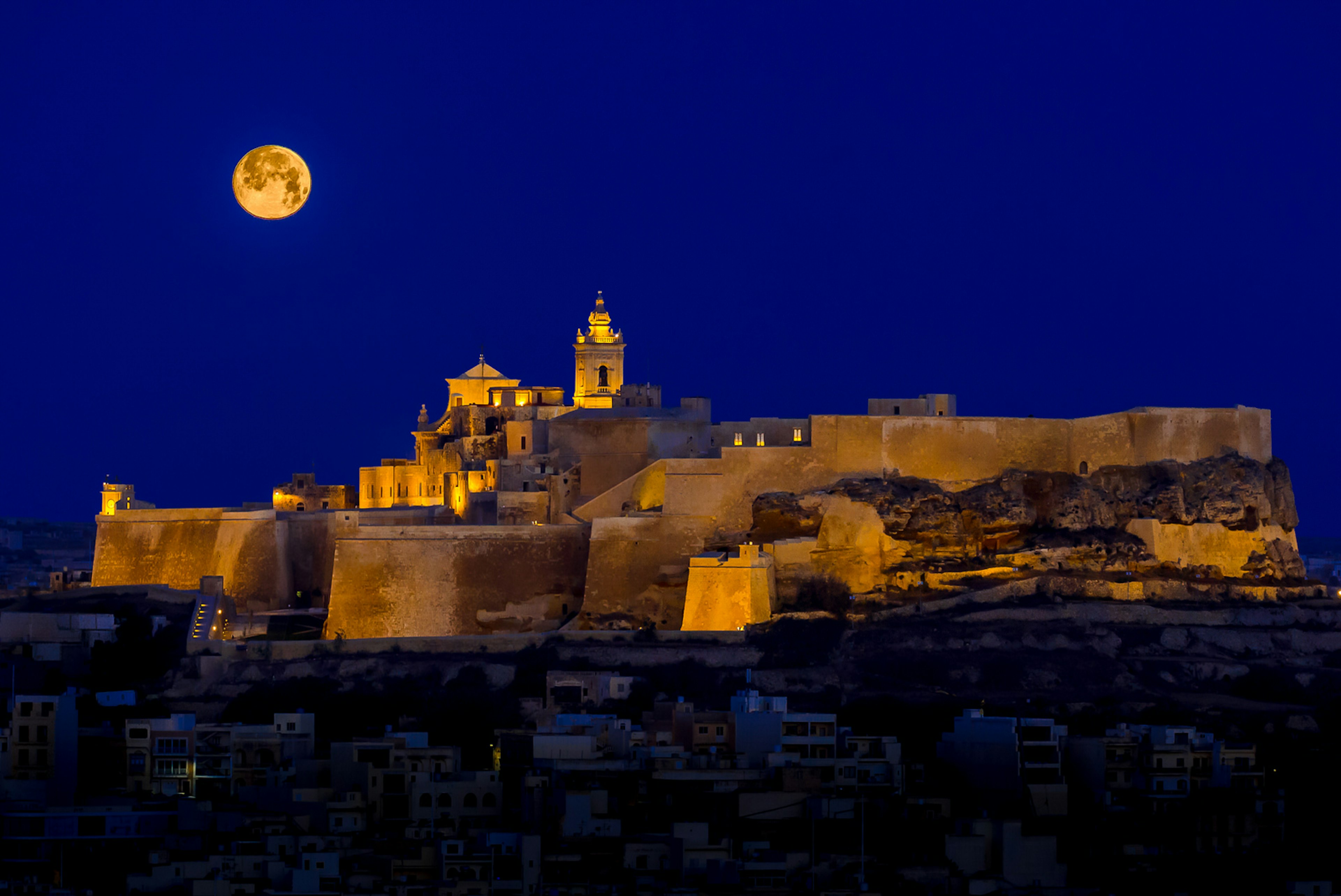 With a full moon rising into a dark sky, the Ċittadella glows gold in floodlights © rossmagri / Getty Images