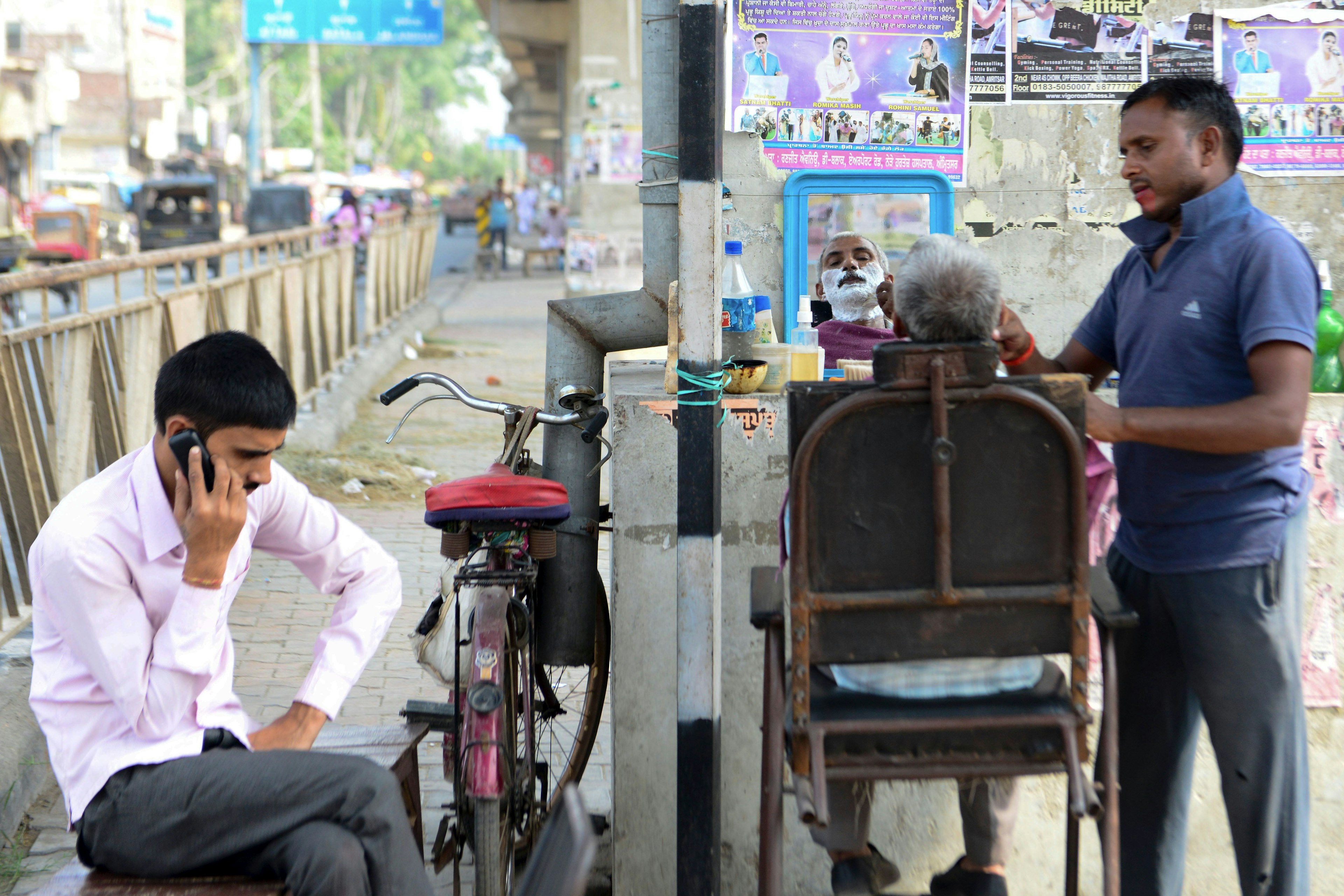 Outside, a man is sitting in a reclining chair while a barber adds shaving foam to his face. Another man is on his mobile phone as he waits his turn.