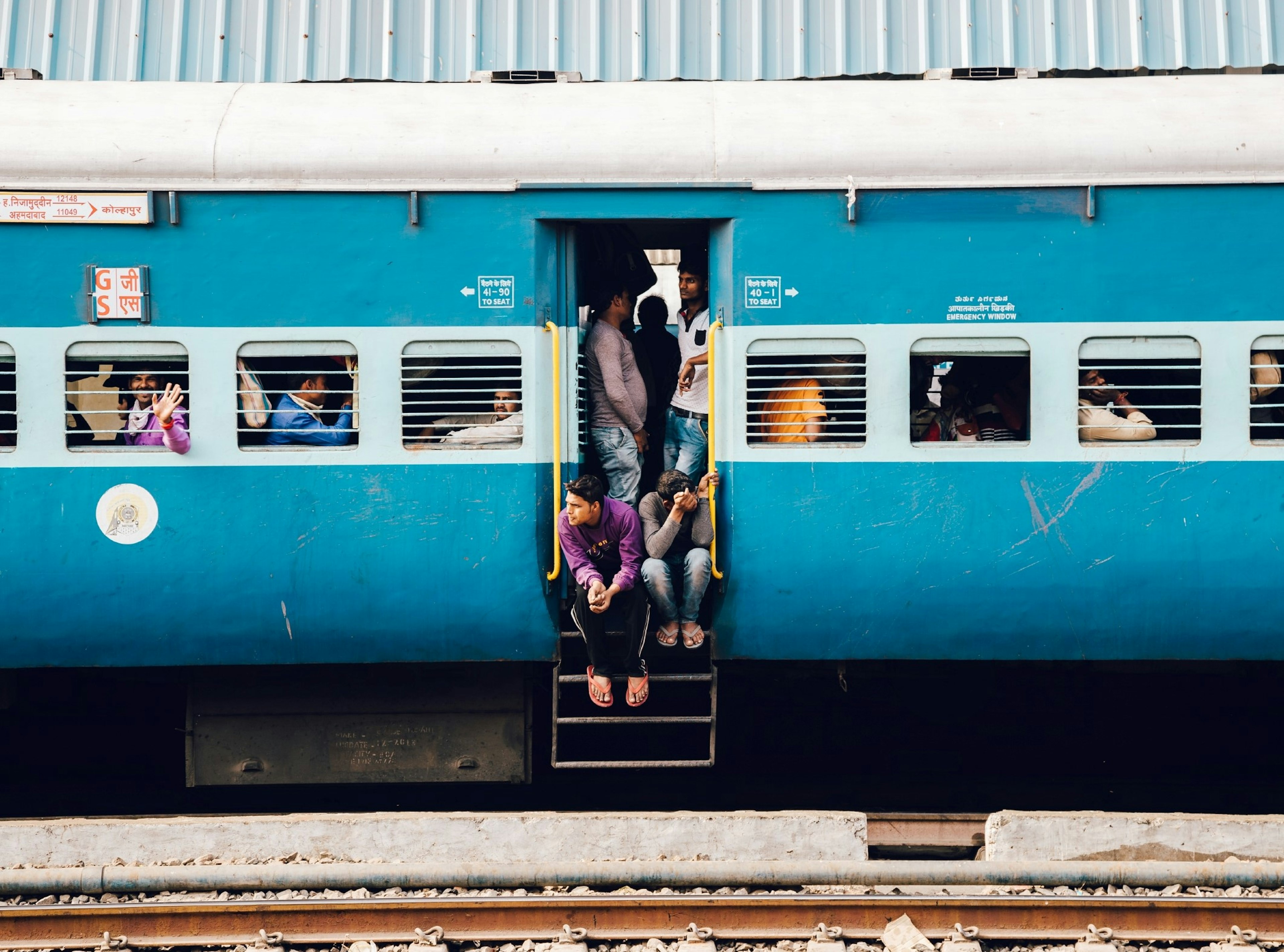 A stationary blue train in India, with passengers sitting and leaning out of the doorway and looking through the train's windows.