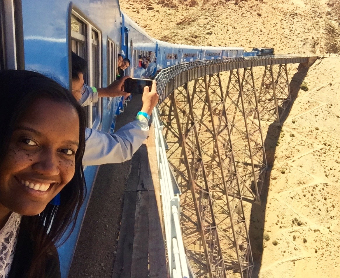 A woman with her head out of a train window smiles at the camera. In the background a number of people are also sticking their heads out of the train's windows to take selfies