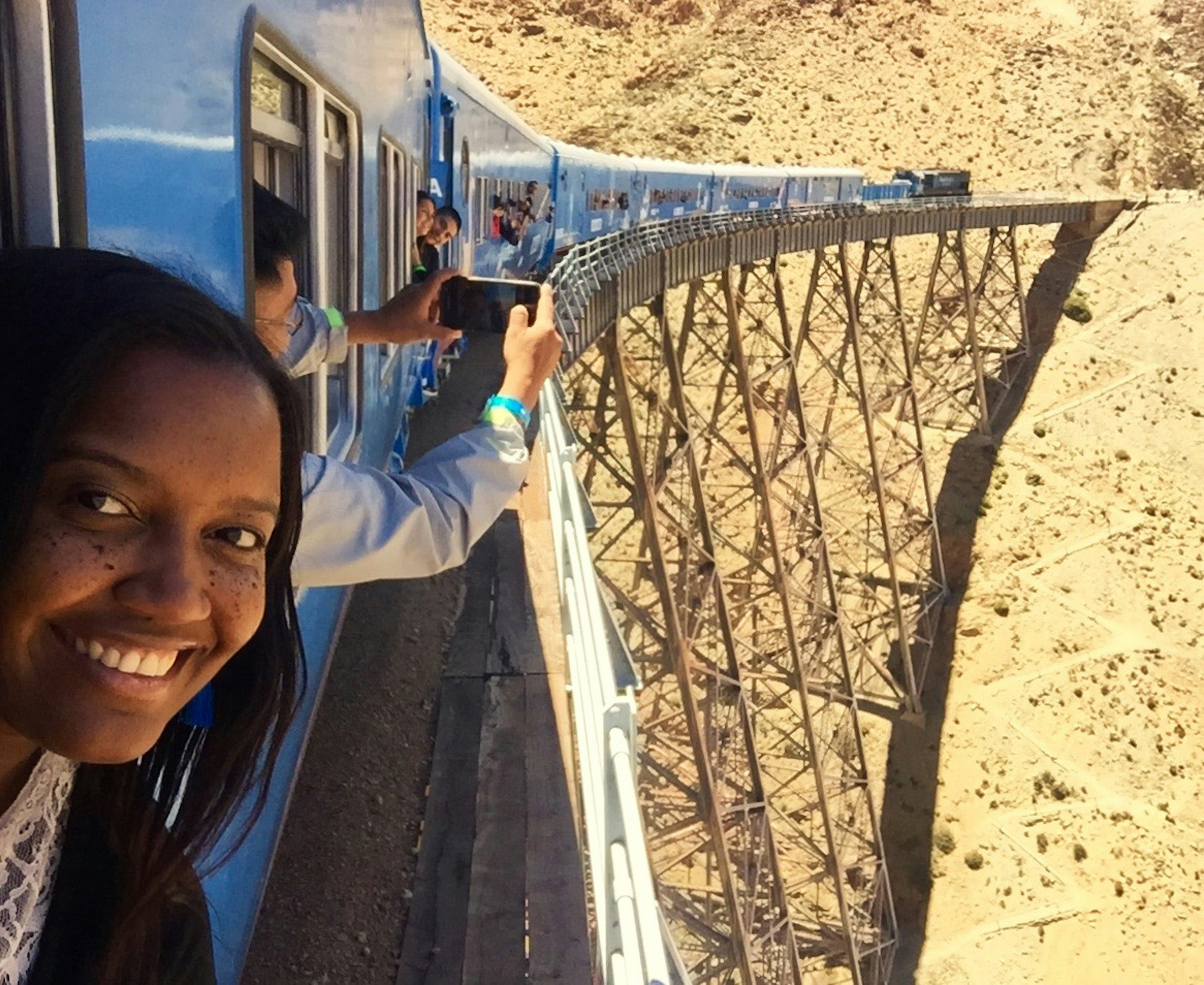 A woman with her head out of a train window smiles at the camera. In the background a number of people are also sticking their heads out of the train's windows to take selfies