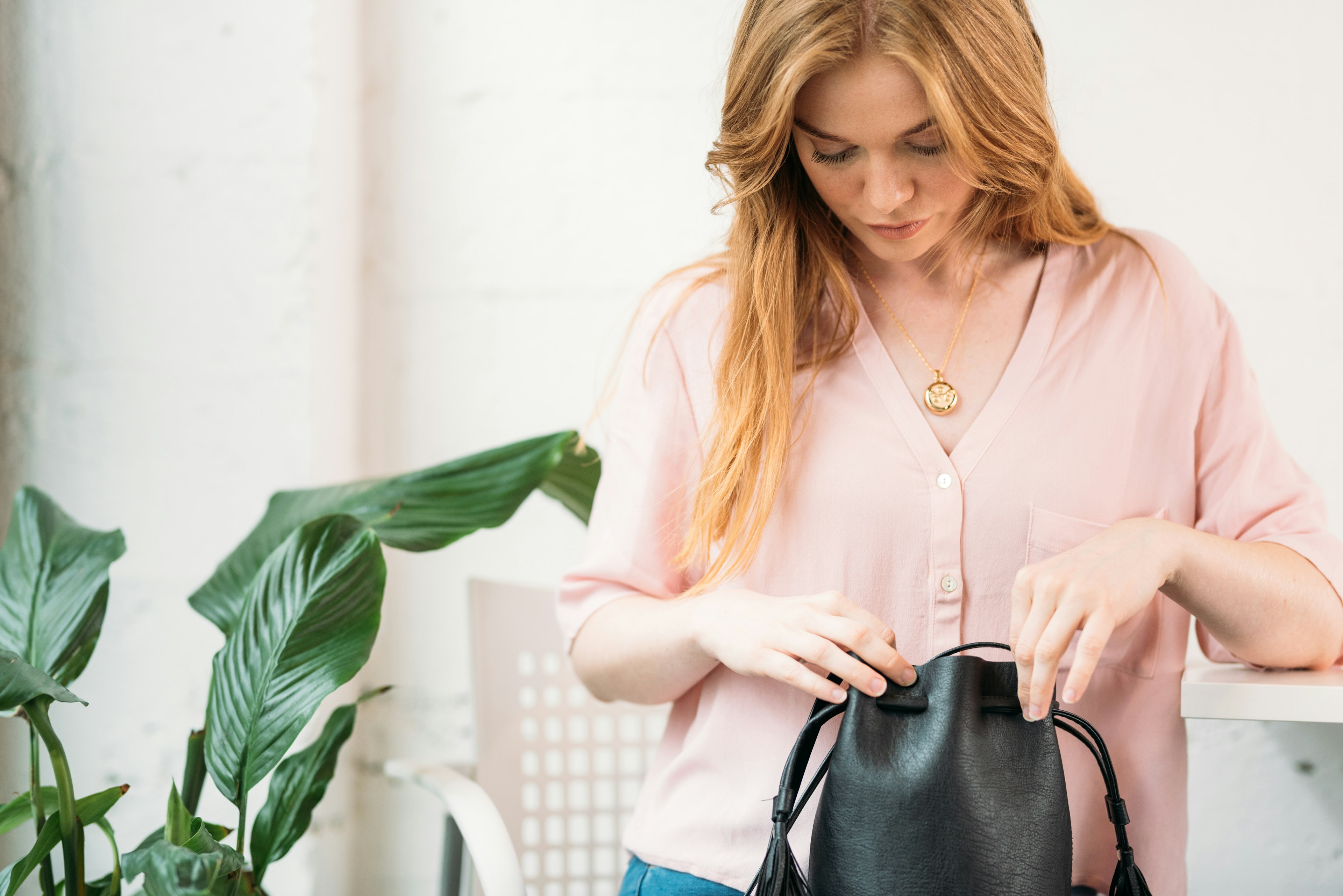 A woman in a pink blouse is looking down into her black handbag; she is wearing a gold invisaWear necklace.