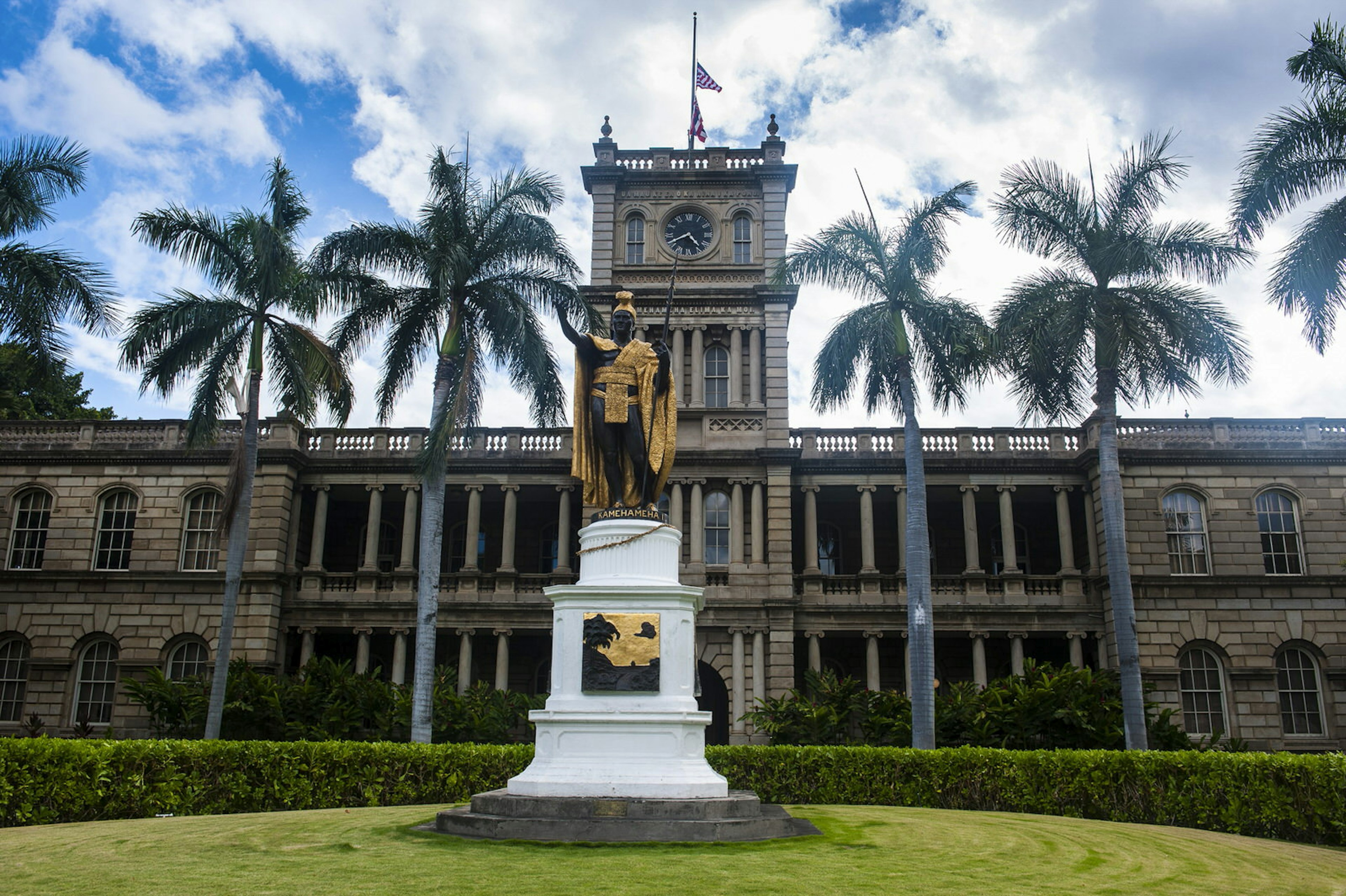 The palace is seen in the background, with a row of palm trees in the middle and a black statue on a white pedestal coverd in gilded traditional robes in the foreground © Michael Runkel / Getty Images