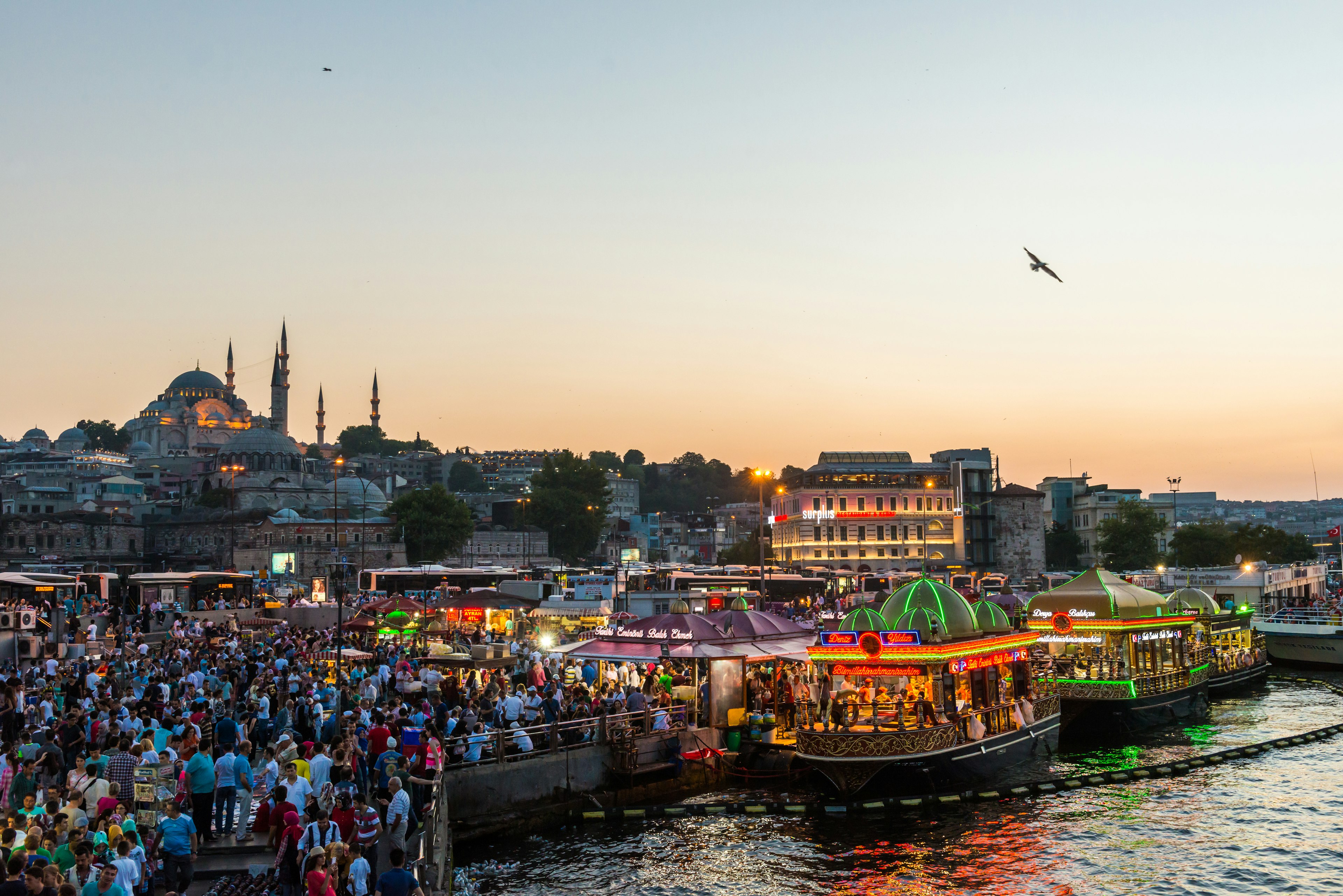 A large group of people walk along the Golden Horn in Istanabul at sunset. There are large brightly lit boats docked on the harbour.