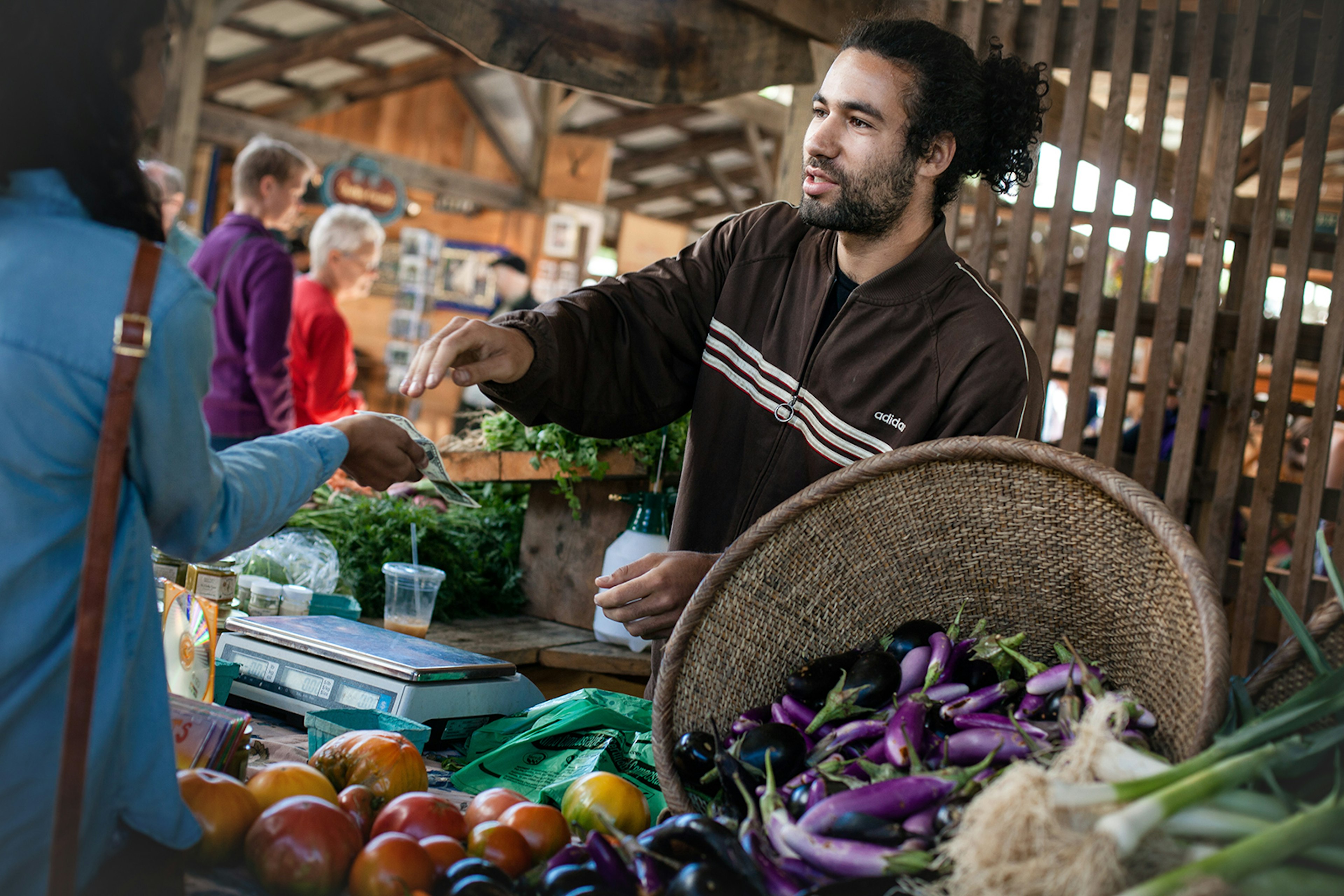 A man smiles and hands change to a woman over a stall of fresh local vegetables (eggplant, etc) at a farmer's market