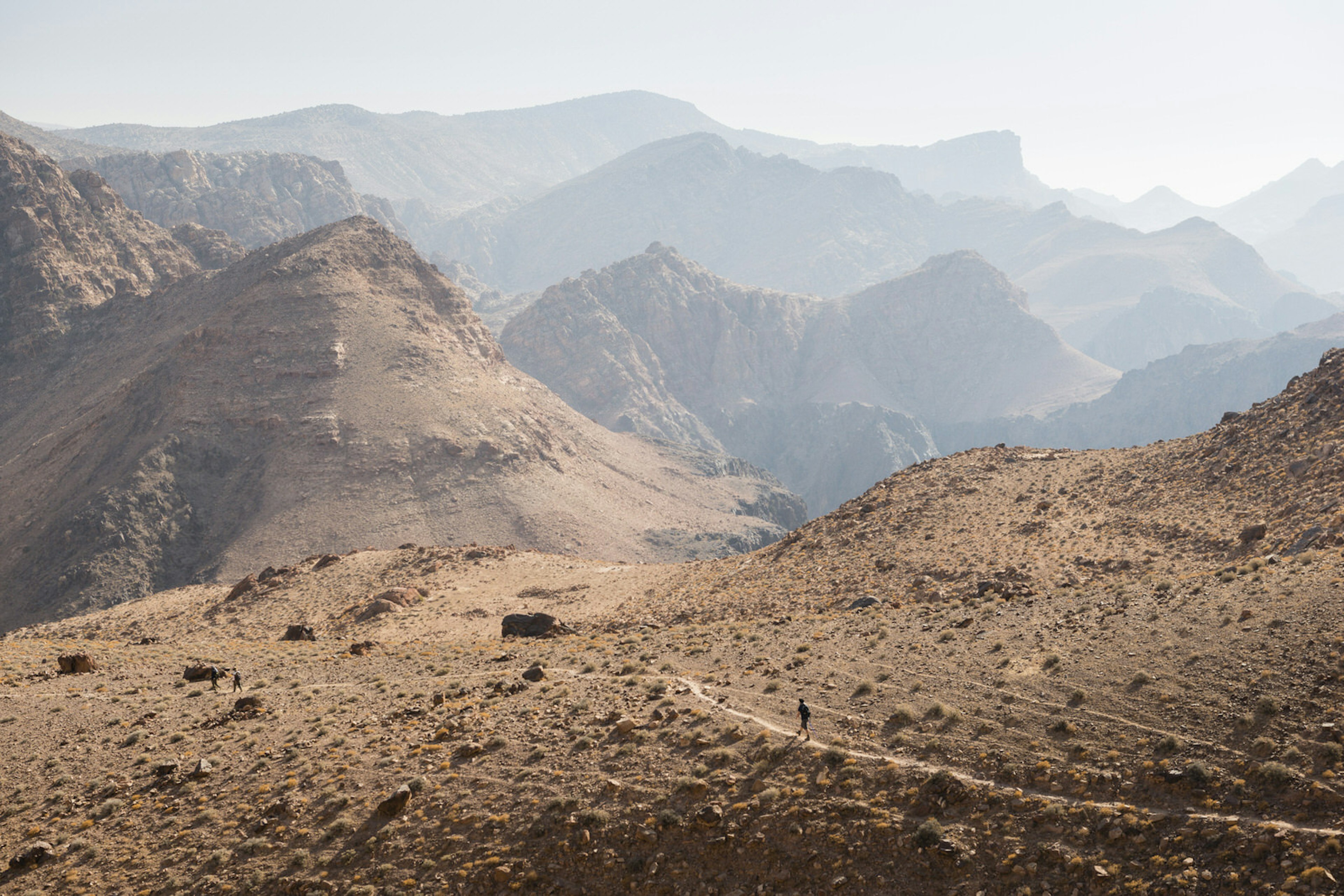 Hikers walk along a small path on the Jordan Trail with numerous mountains visible in the background